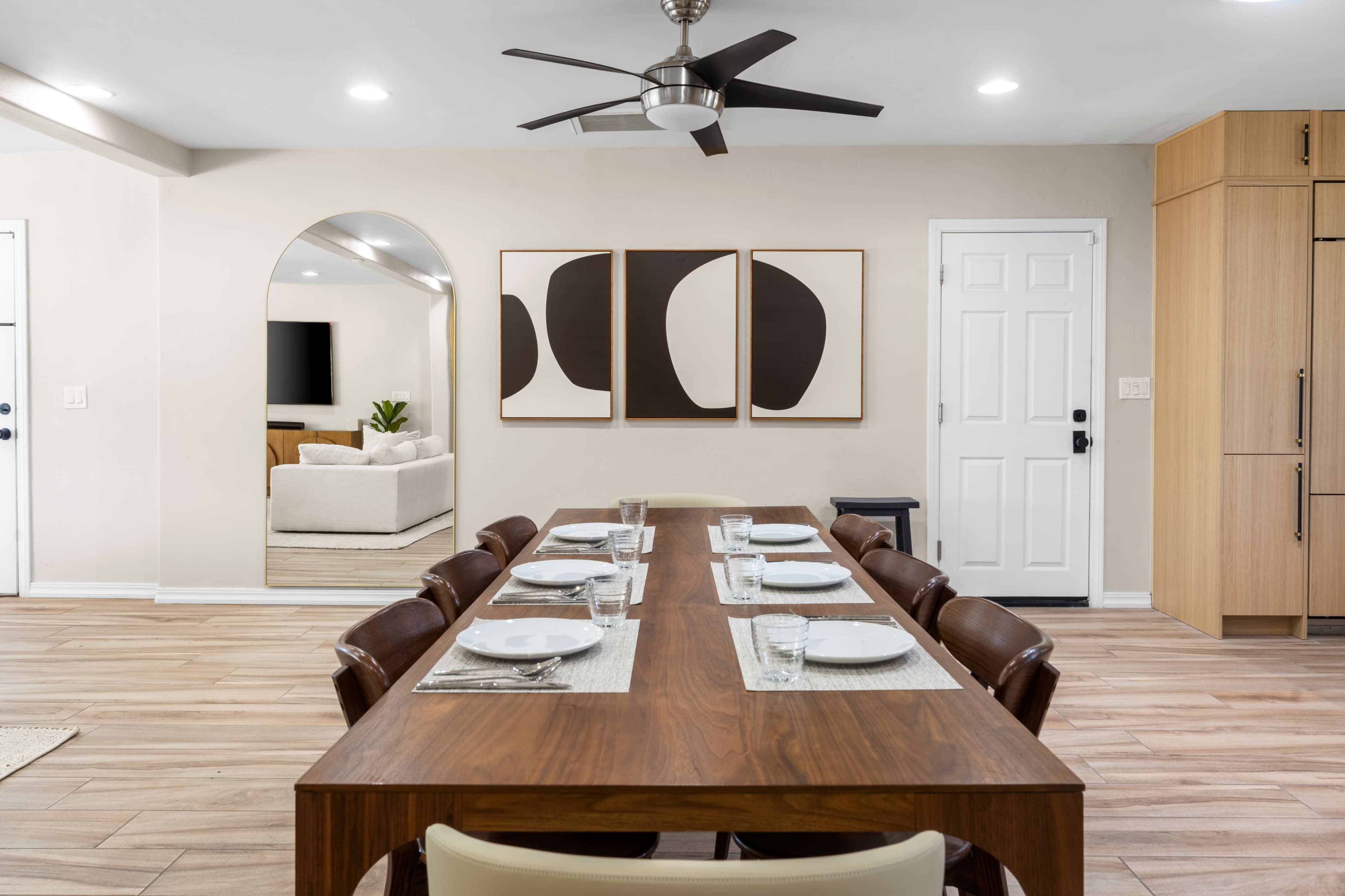 A modern dining area features a long wooden table set for eight, with artwork on the wall and a large ceiling fan overhead.