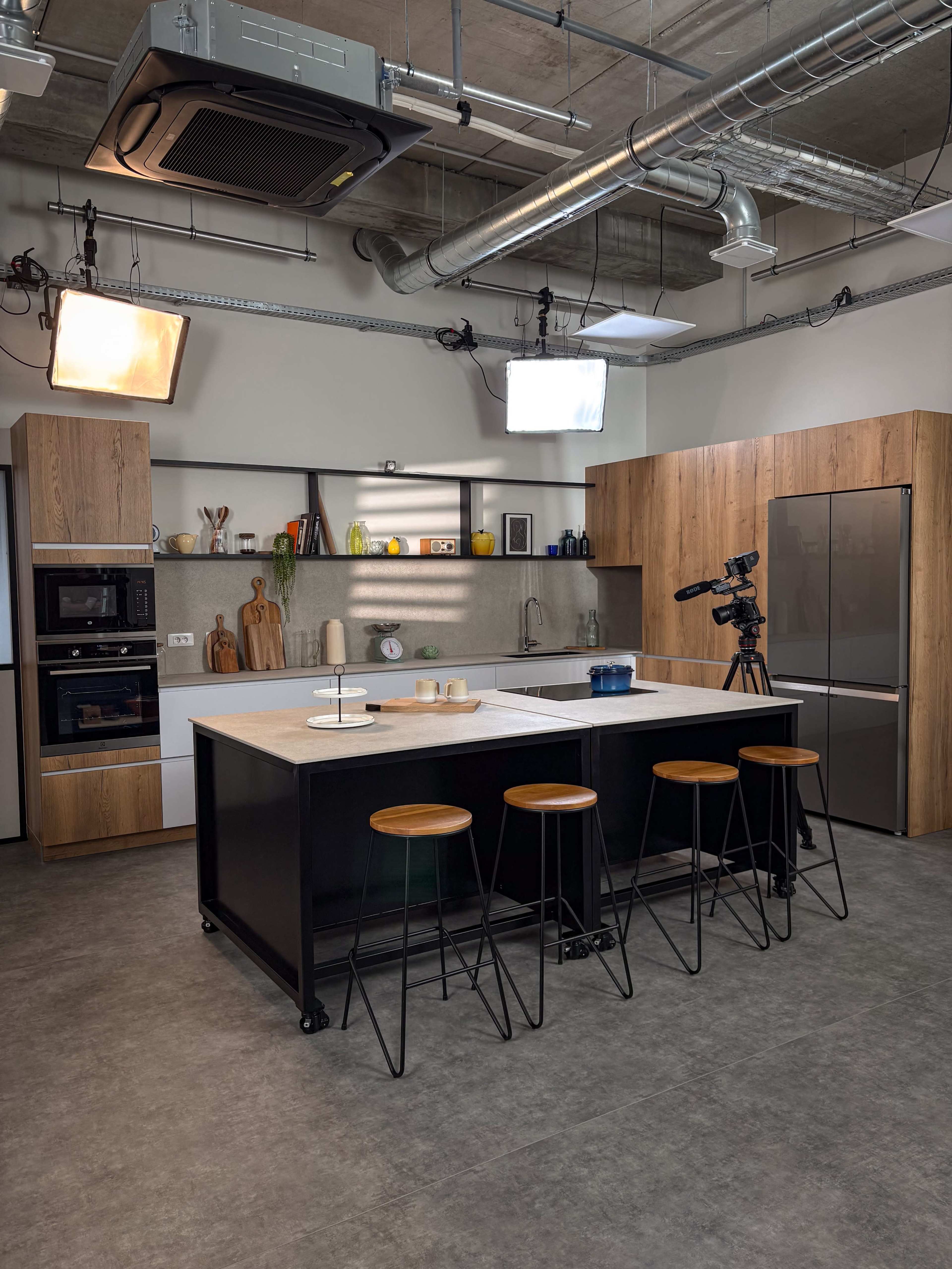 The image shows a modern kitchen setup with wooden cabinets, a center island, bar stools, and a camera positioned nearby for filming.