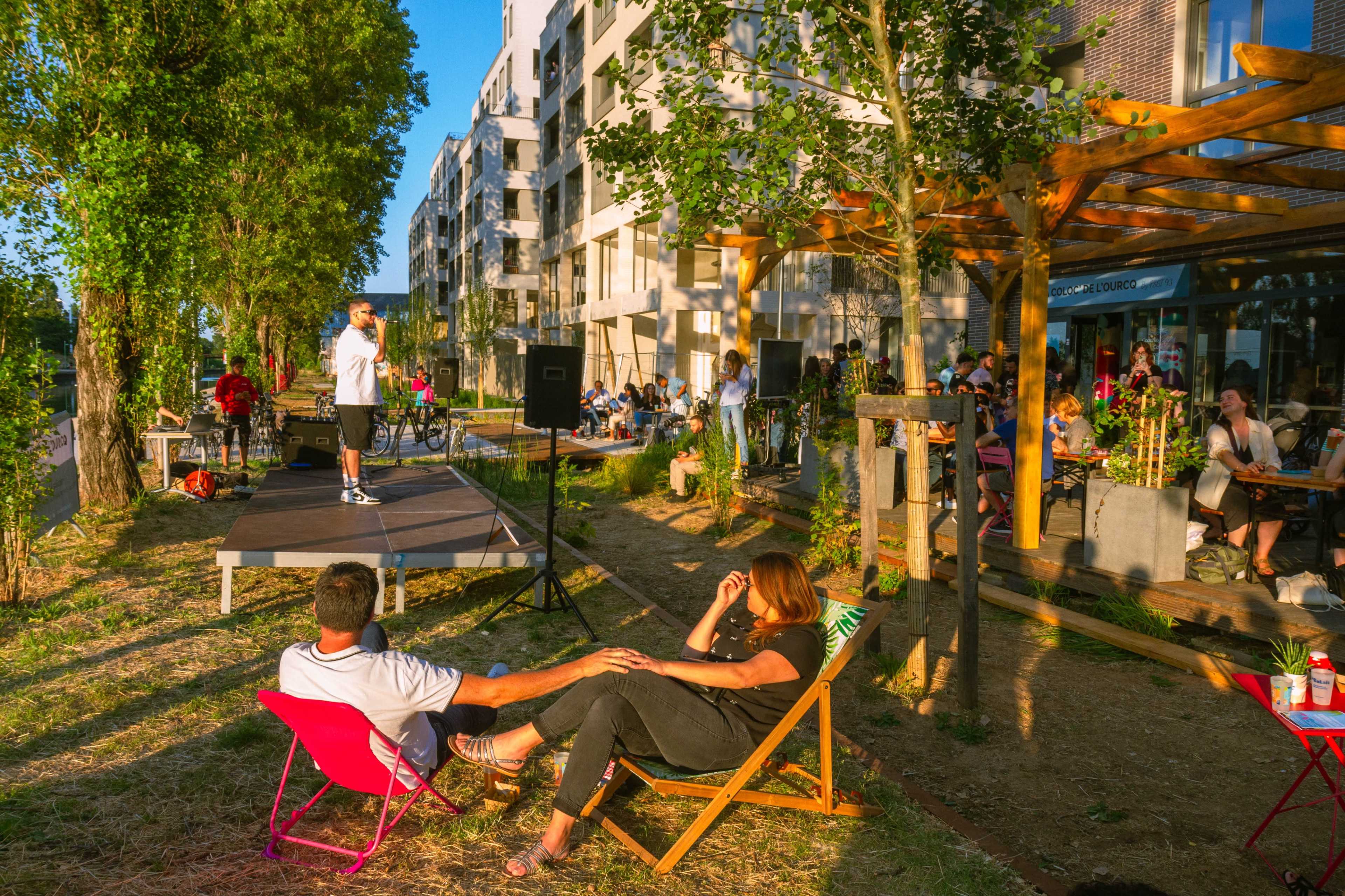 A performer stands on a stage in a green space while an audience of seated people relaxes on lawn chairs and tables nearby, surrounded by modern apartment buildings.
