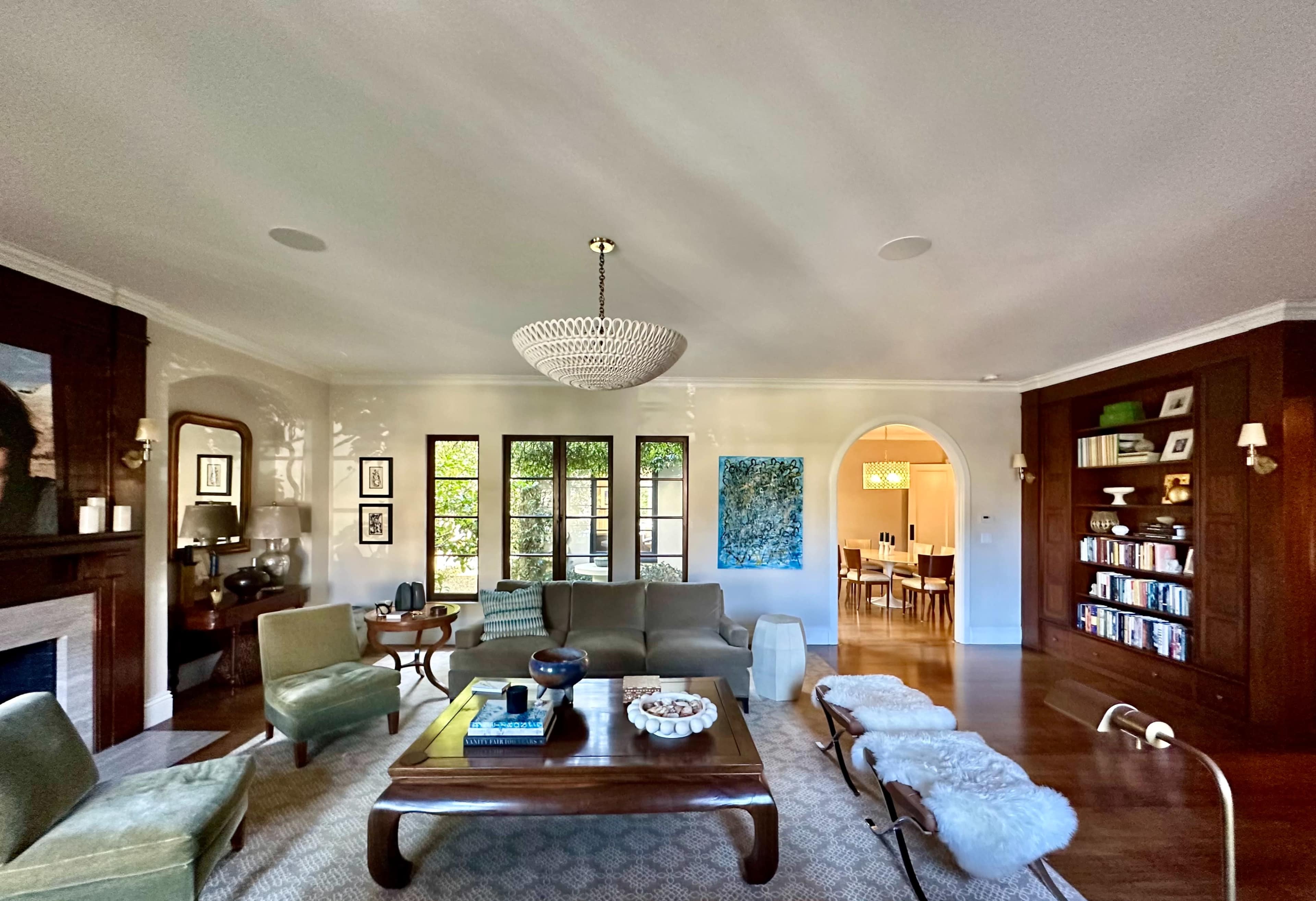 The image shows a spacious living room with a chandelier, a green sofa, and a coffee table, alongside large windows and a doorway leading to another room.
