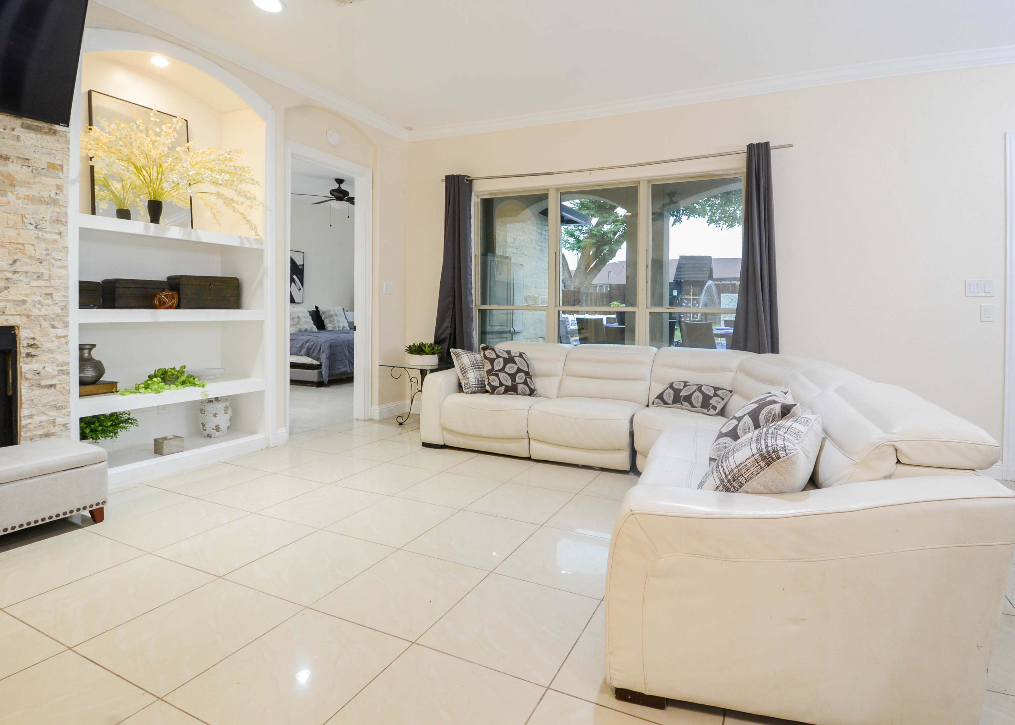 The image features a spacious living room with a white sectional sofa, large windows, and a built-in bookshelf displaying decorative items.