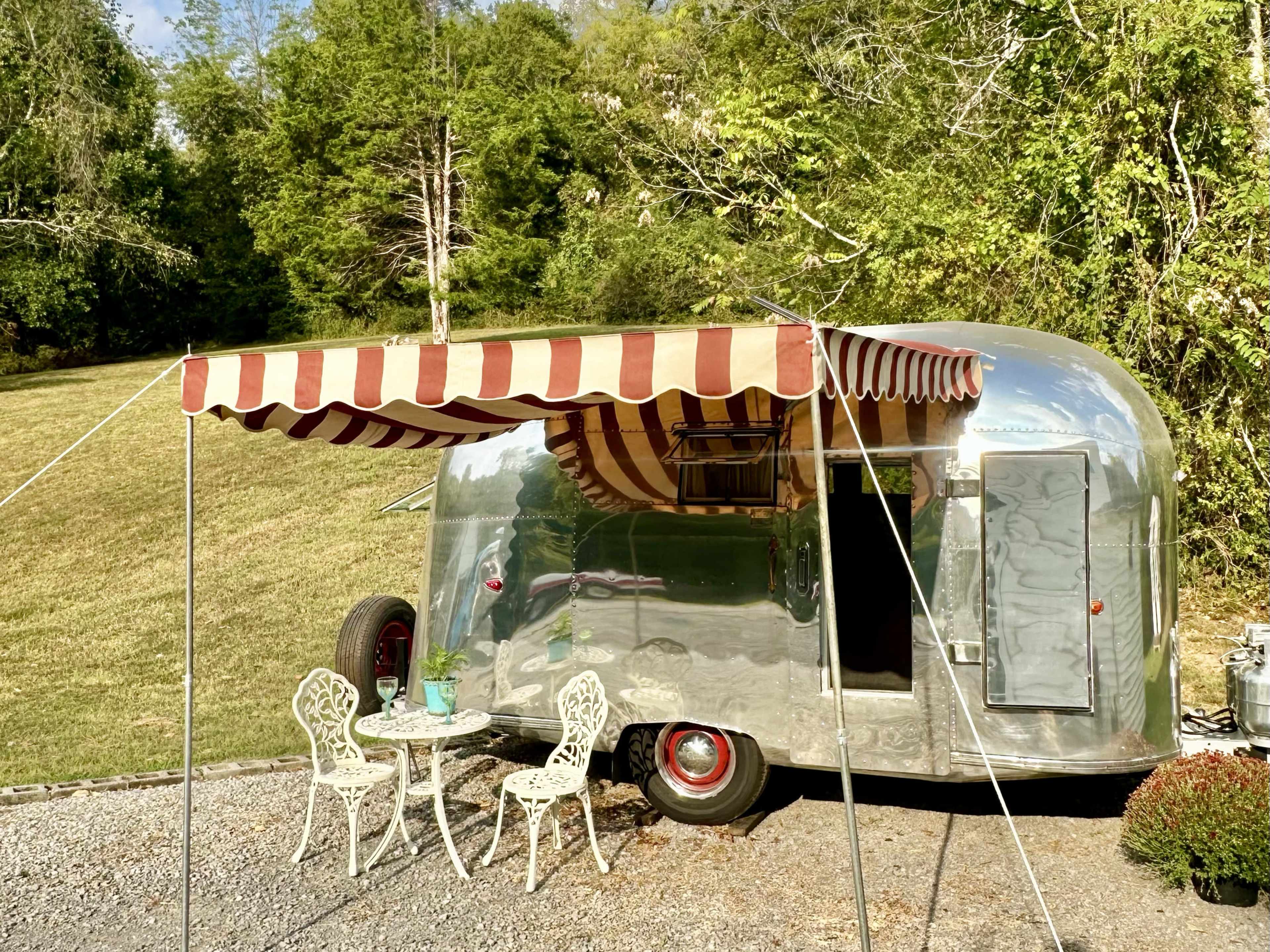 The image shows a vintage silver trailer with a striped awning, set up next to a small outdoor table and chairs on a gravel surface, surrounded by greenery.