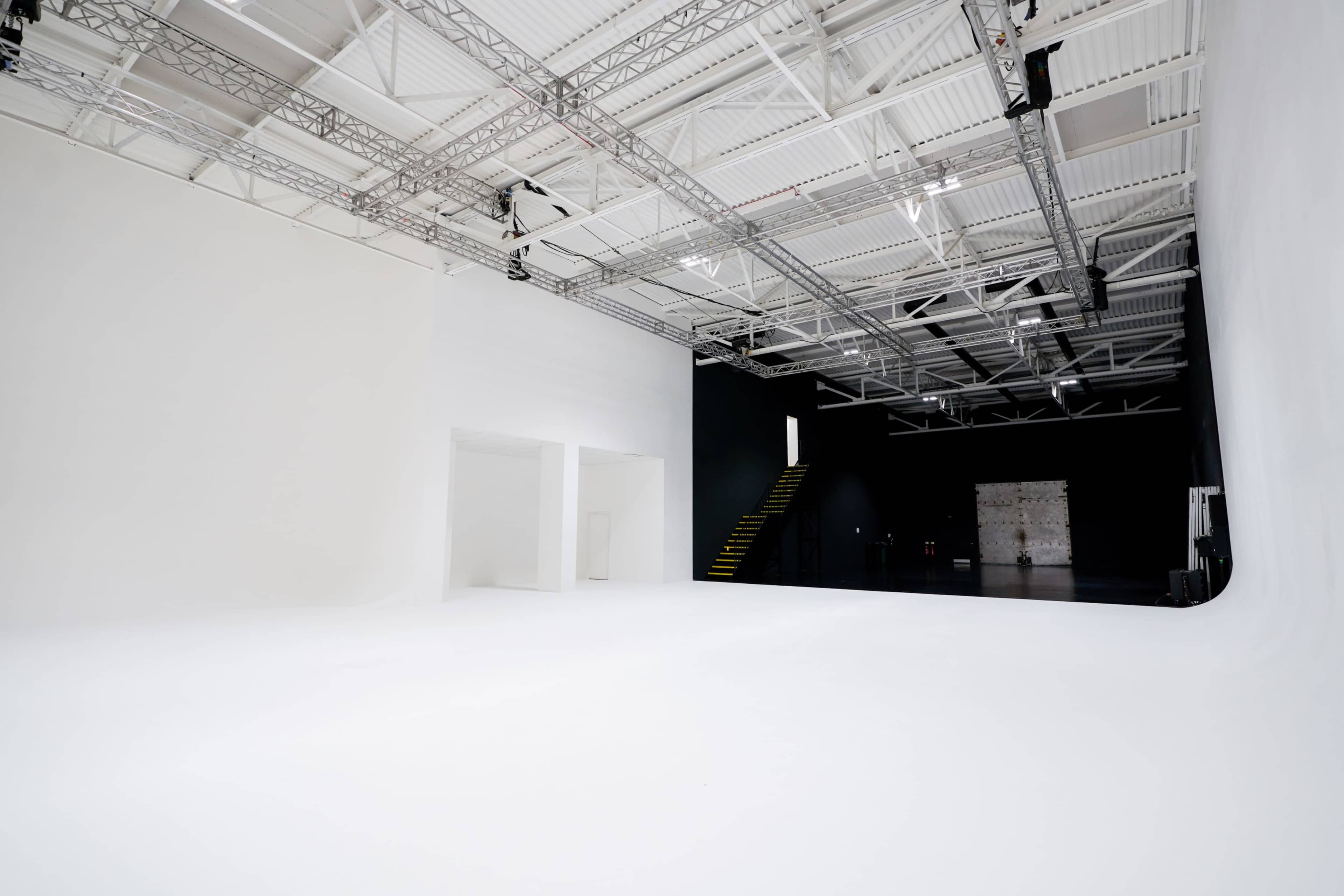 The image shows a spacious studio with a stark white backdrop, contrasting with a dark wall on one side and visible lighting fixtures overhead.