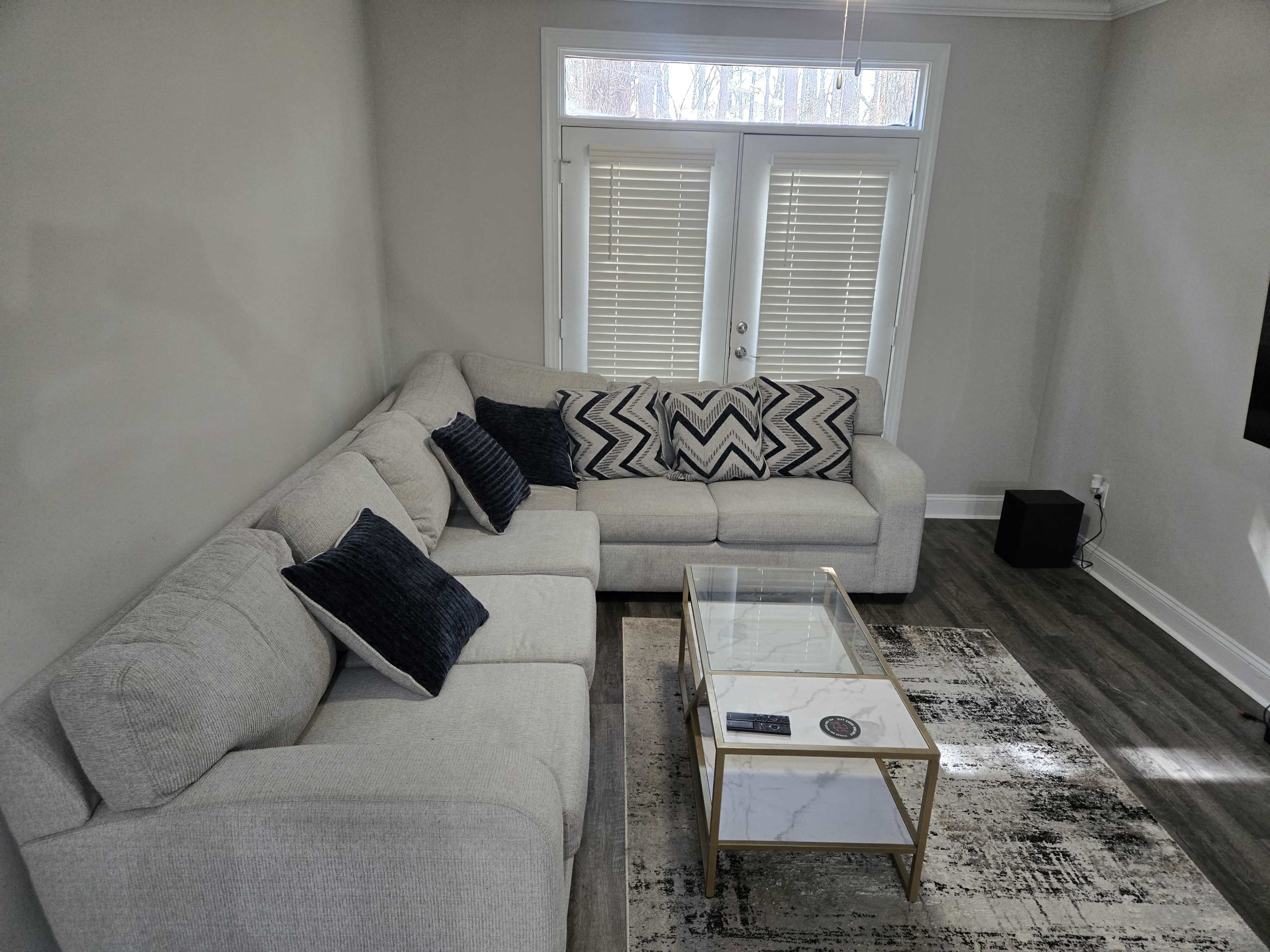 The image shows a light-colored living room with a large sectional couch, a glass coffee table, and a patterned area rug.