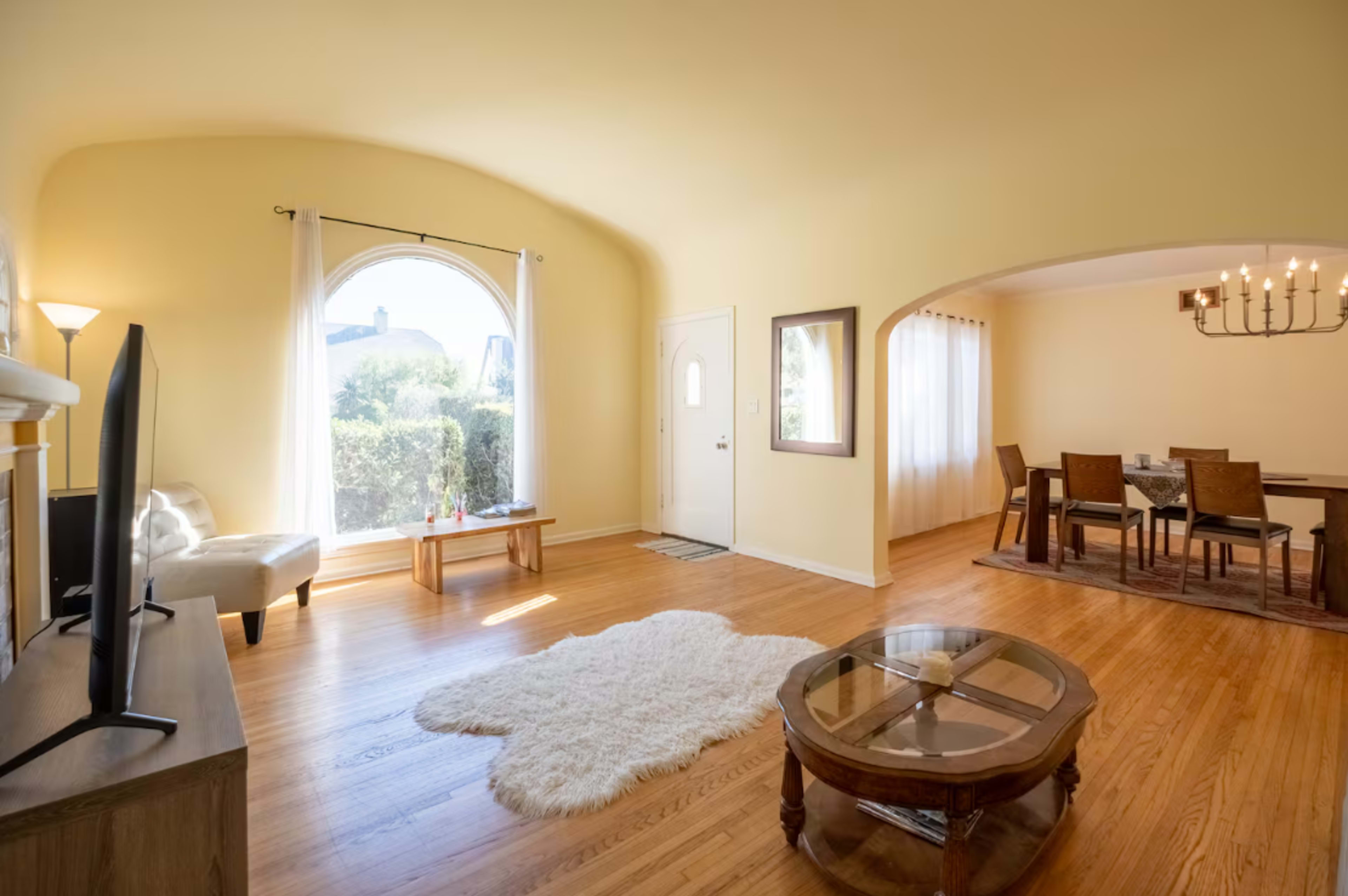 The image shows a spacious living area with hardwood floors, a large window, and a dining area with a table and chairs.