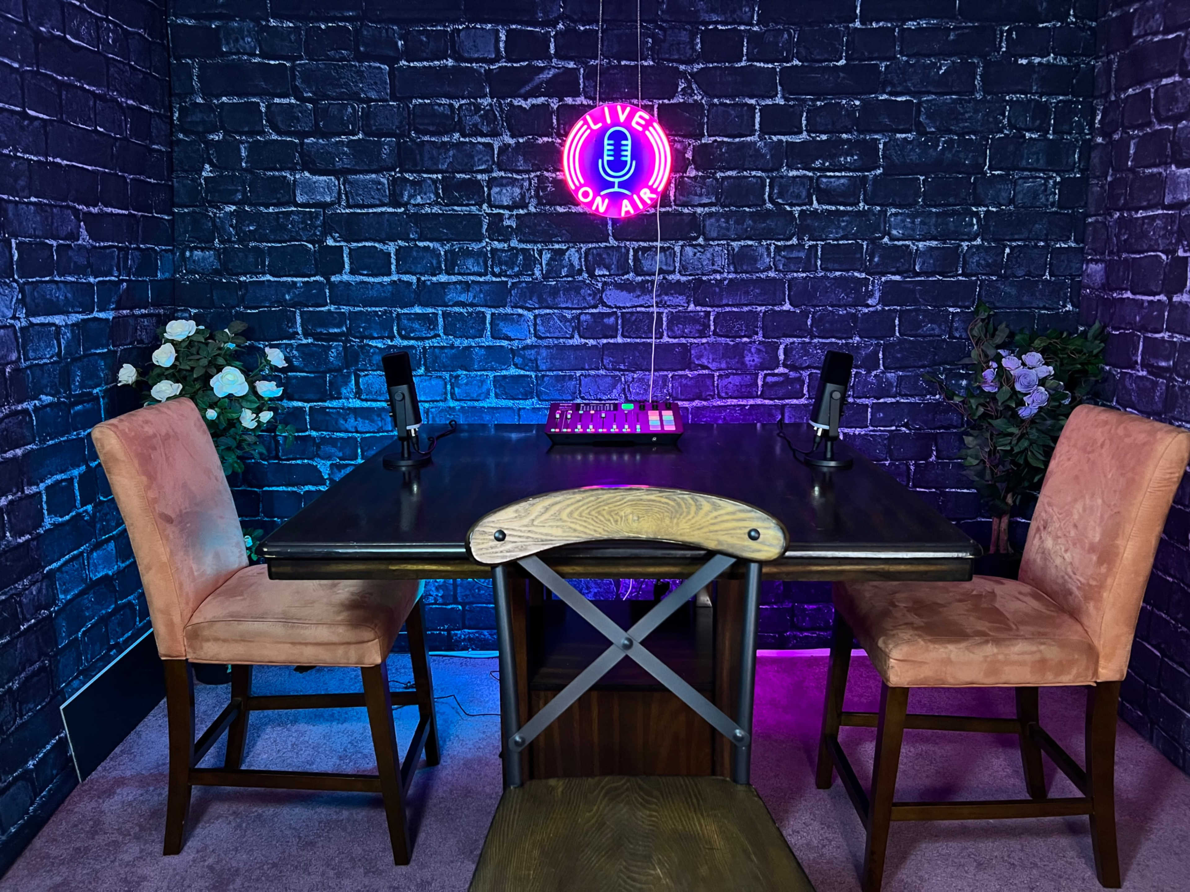 A dark brick wall serves as the backdrop for a small table with two microphones and two chairs, illuminated by a neon "LIVE ON AIR" sign.