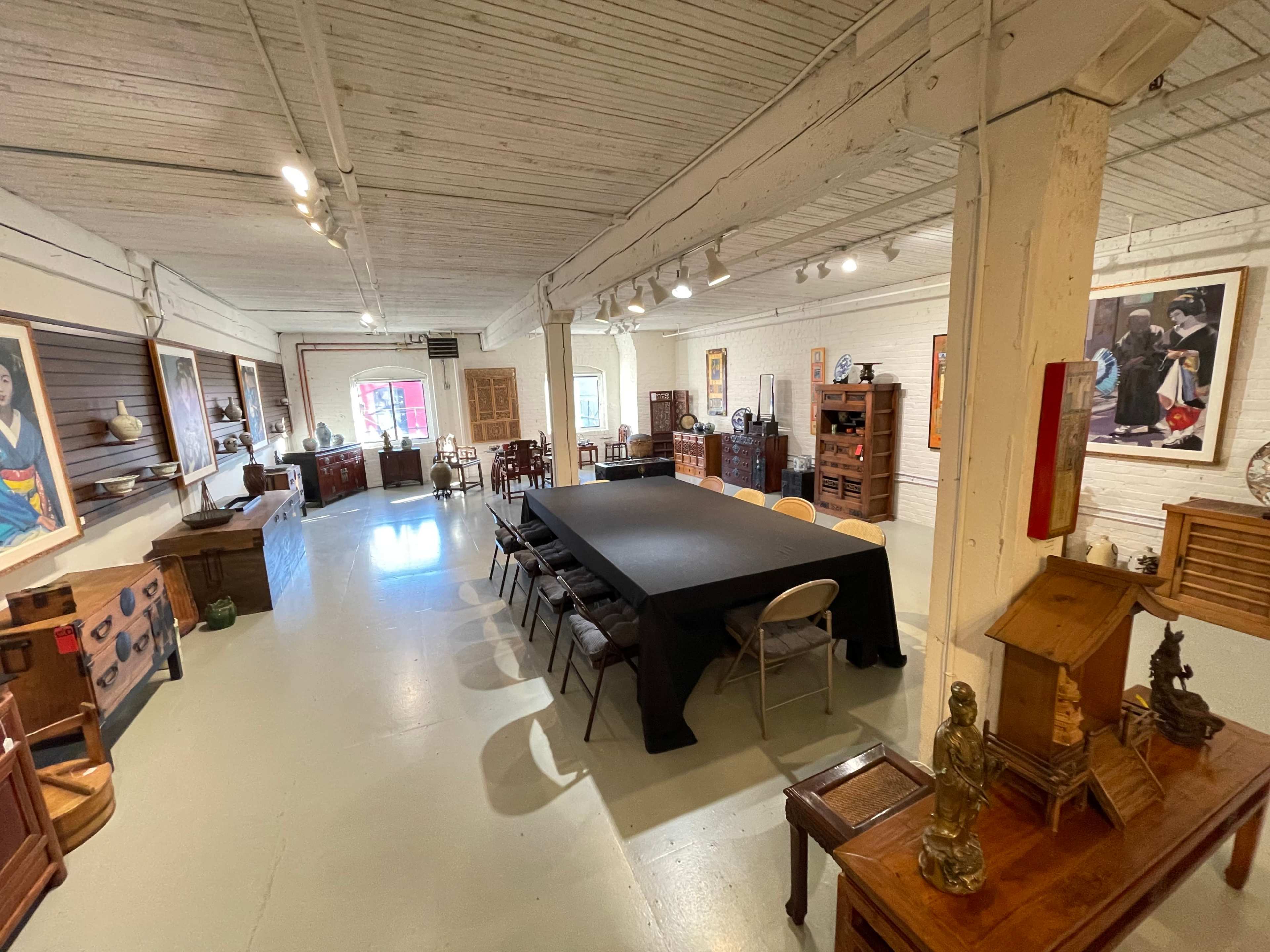 A spacious gallery room with a large black table surrounded by chairs, featuring various wooden furniture and artwork displayed on the walls.