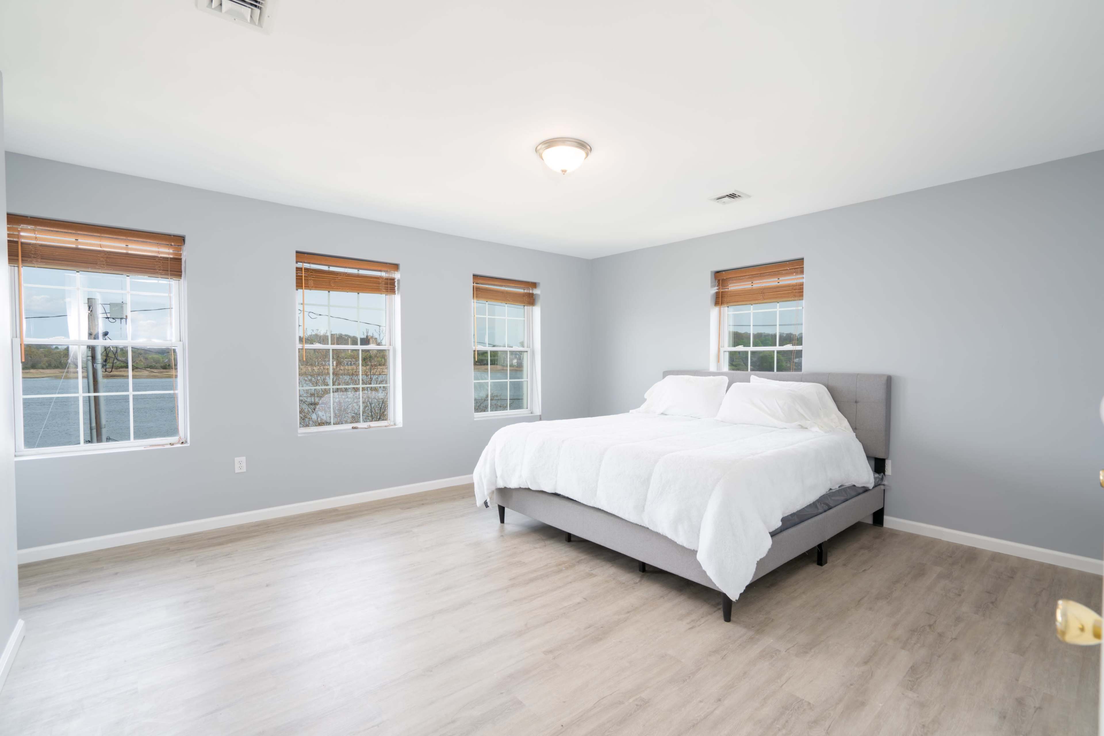 A minimalist bedroom features a bed with white bedding and four windows with wooden blinds.