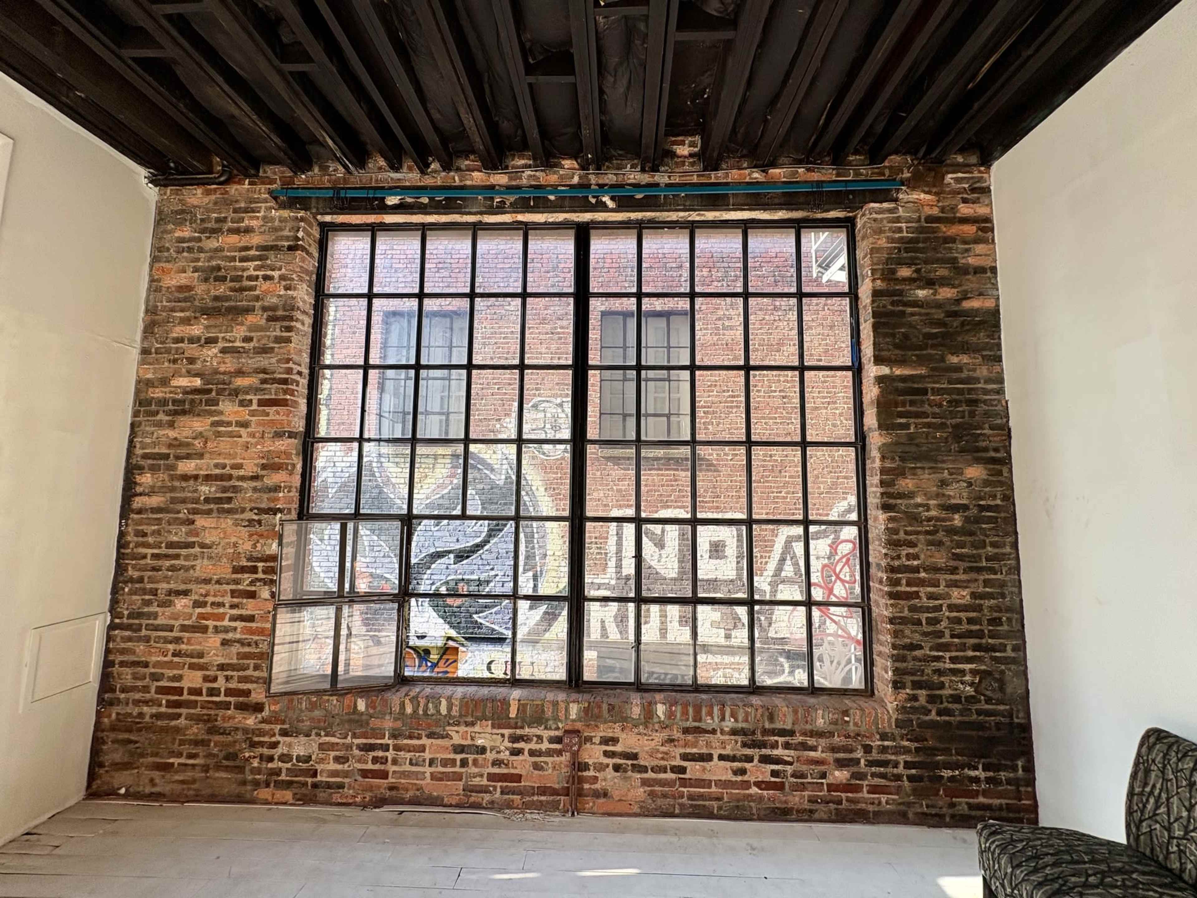 The image shows an empty interior space with a large industrial window showcasing a brick wall and graffiti on the opposite side.