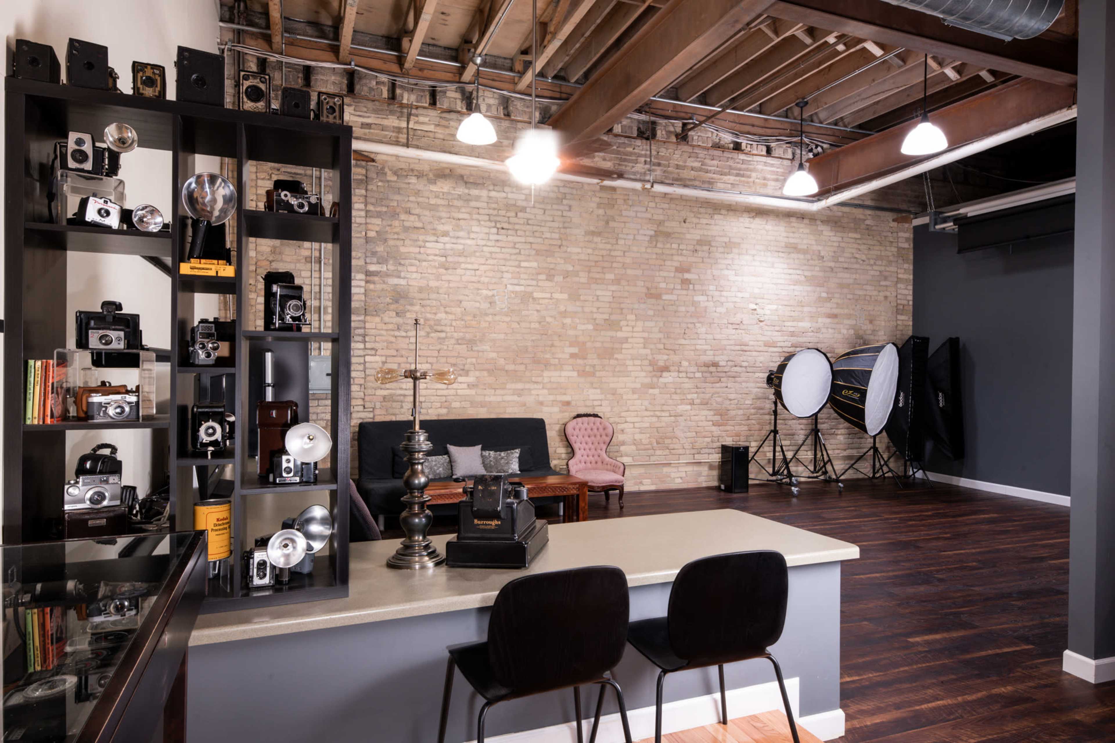 A spacious photography studio with a brick wall, decorative cameras on a black shelf, a seating area with a vintage chair, and large lighting equipment in the background.