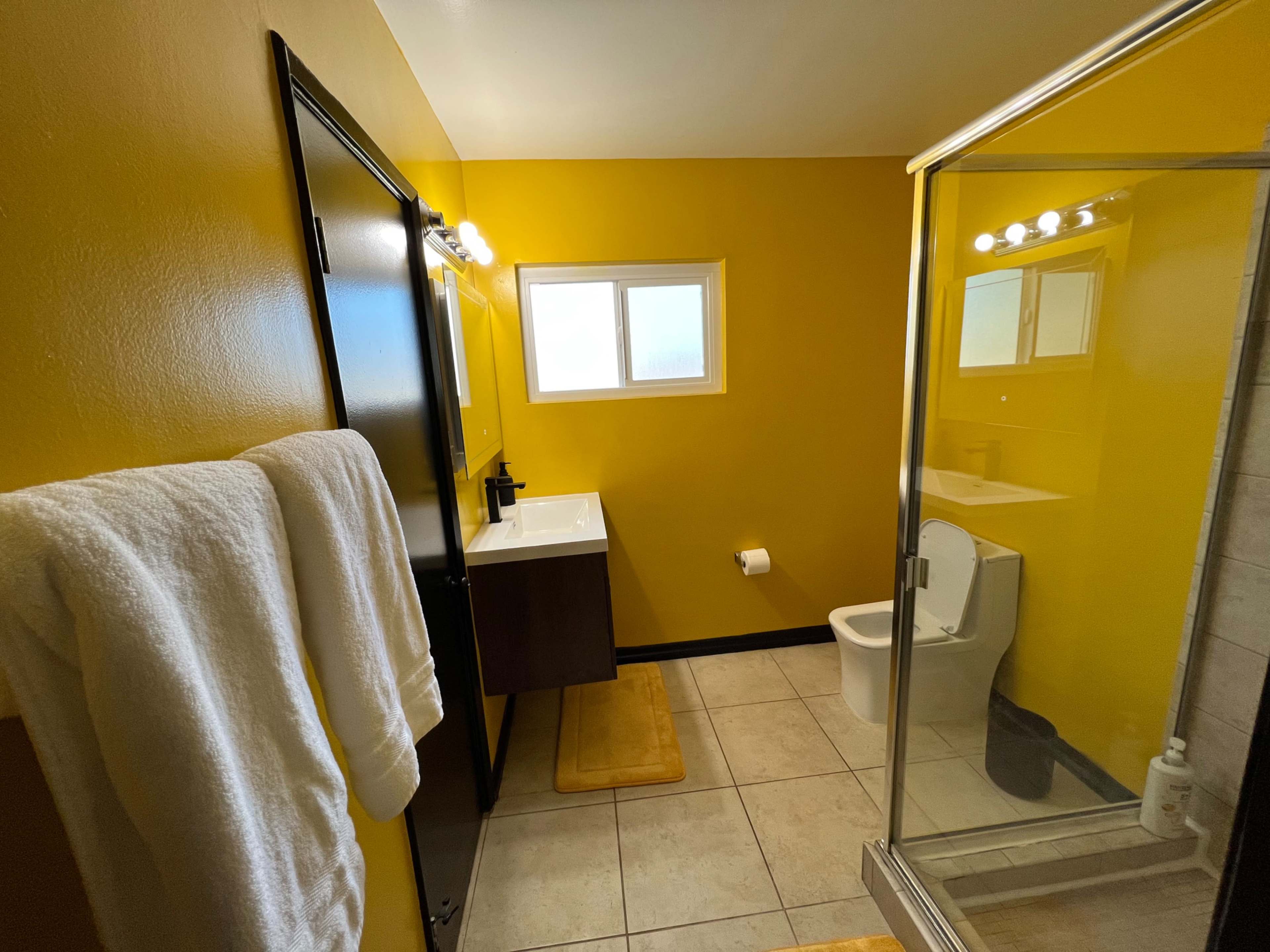 A bathroom featuring a yellow wall, a glass shower enclosure, a toilet, a sink with a mirror, and towels hanging on the wall.