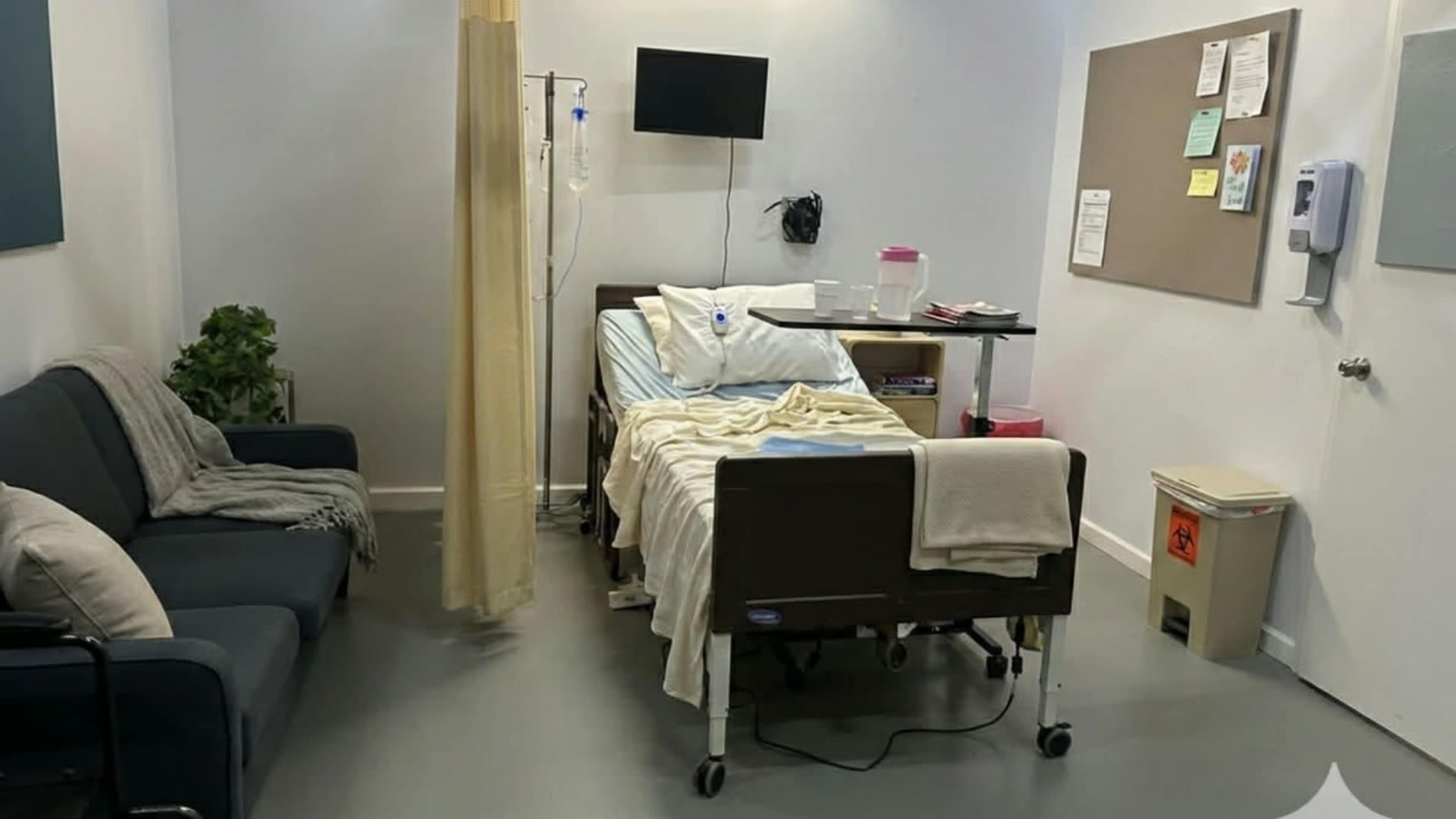 The image shows a hospital room with a bed, an IV stand, a couch, and a bulletin board on the wall.