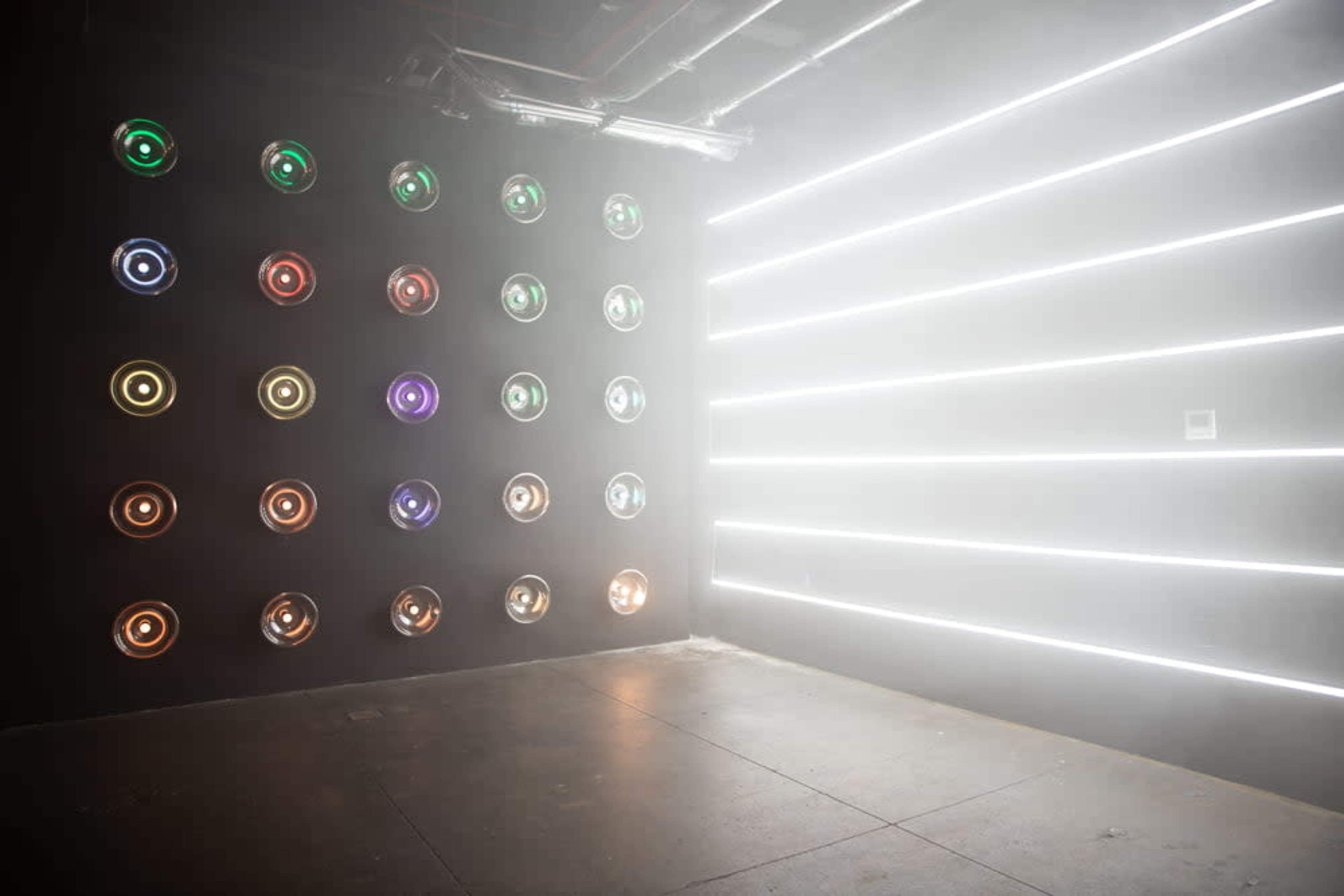 The image shows a dimly lit room with a wall featuring circular light fixtures arranged in a grid pattern, and another wall illuminated by horizontal white light strips amidst a foggy atmosphere.