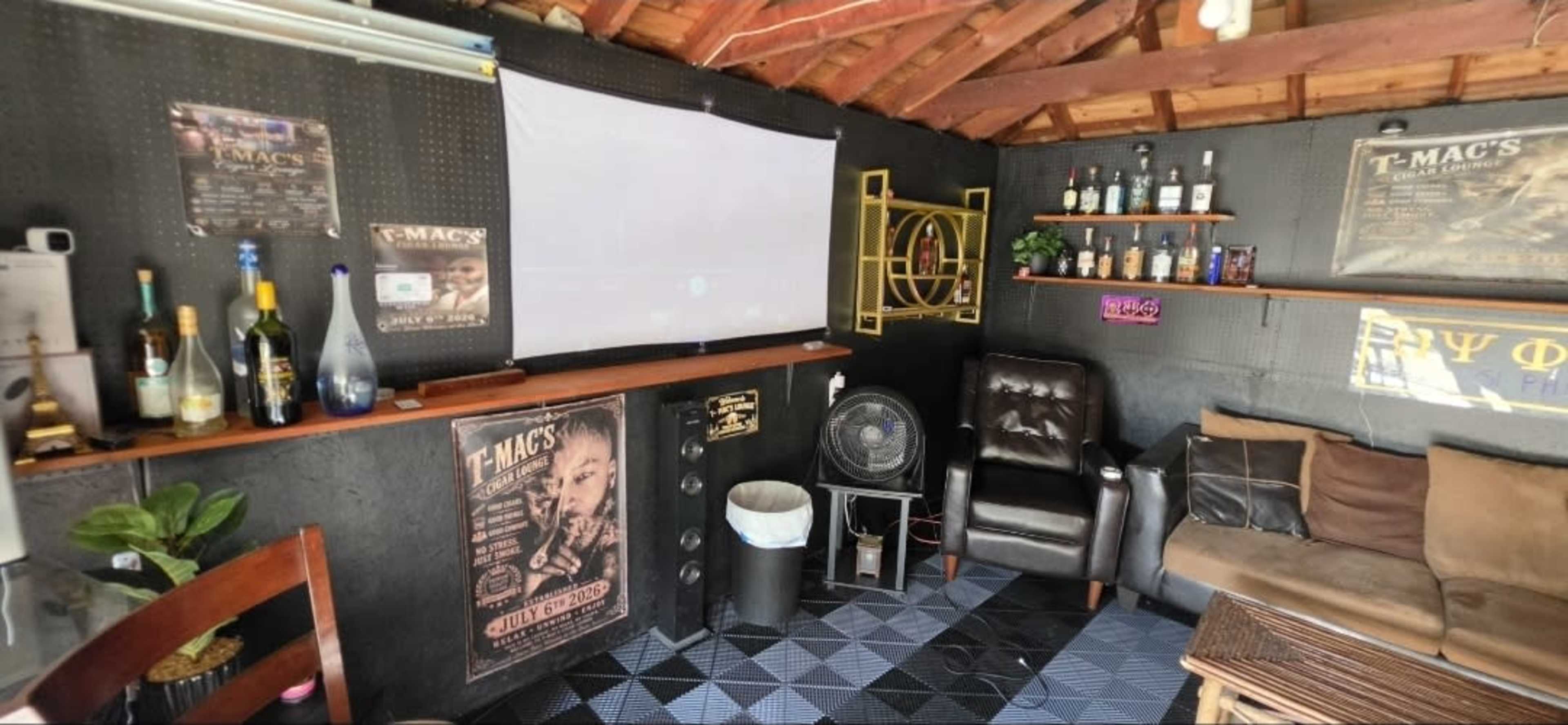 The image shows a small, cozy indoor space featuring a projection screen, a leather chair, a couch, and shelves displaying various bottles and posters on the walls.