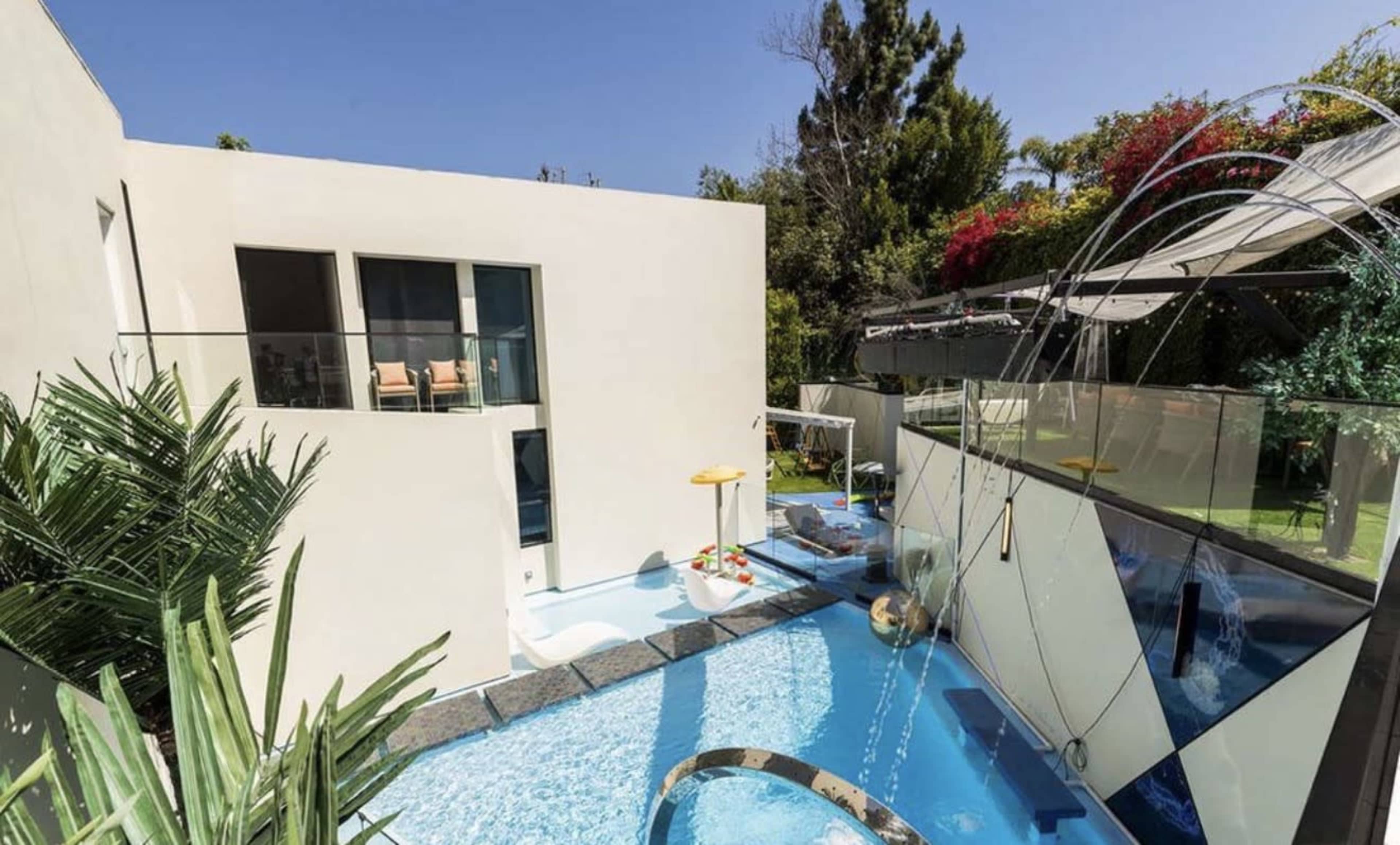 The image shows a modern home with a swimming pool and a patio area surrounded by tropical plants.