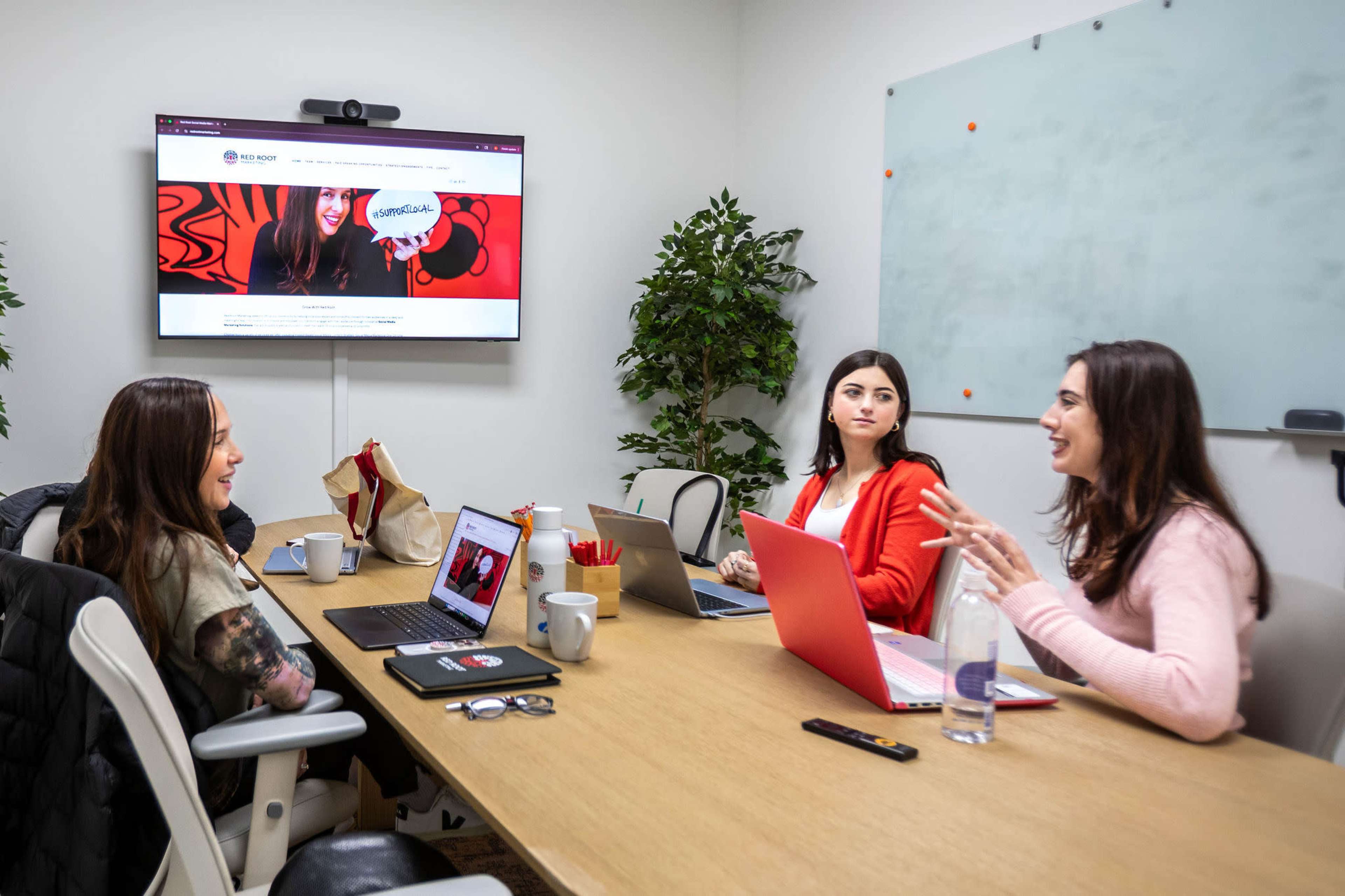 Three women are seated around a conference table with laptops and drinks, engaging in a discussion in a modern meeting room featuring a large screen displaying a website.