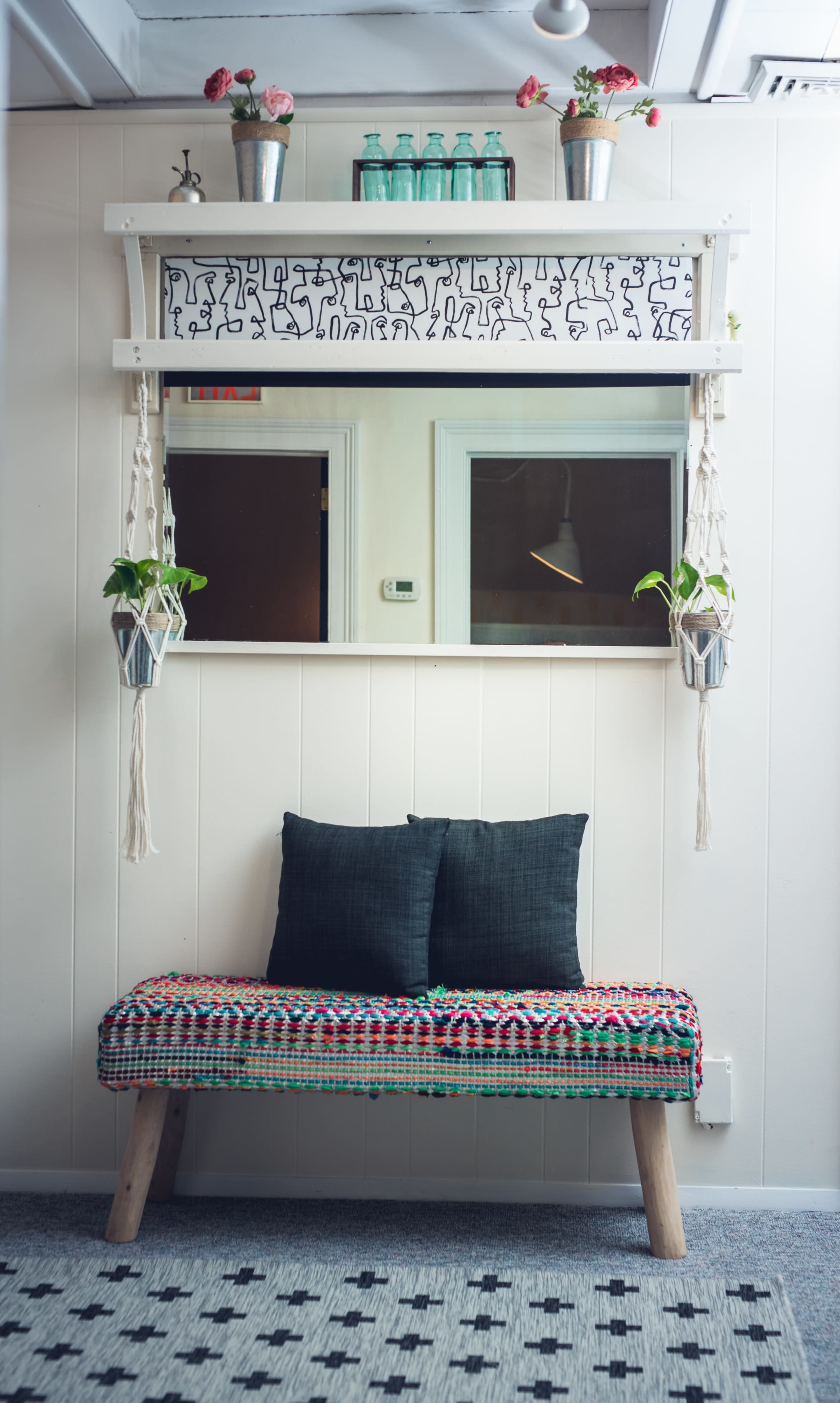 A decorative hallway features a colorful bench with two pillows, a patterned shelf above a mirror, and hanging plants.