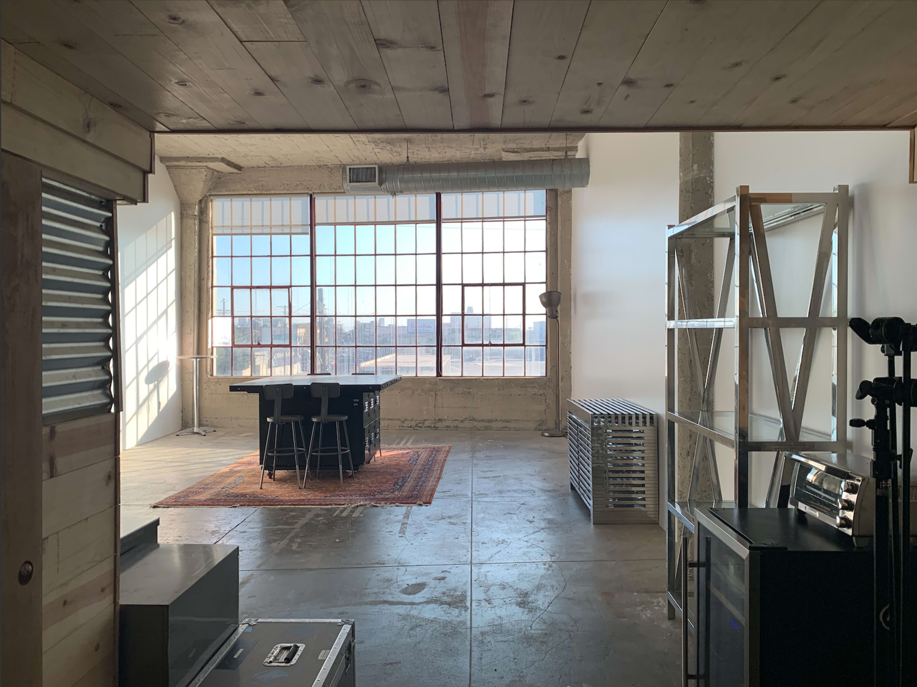 A spacious industrial-style room with large windows, a central table surrounded by chairs, and various equipment and furniture along the walls.
