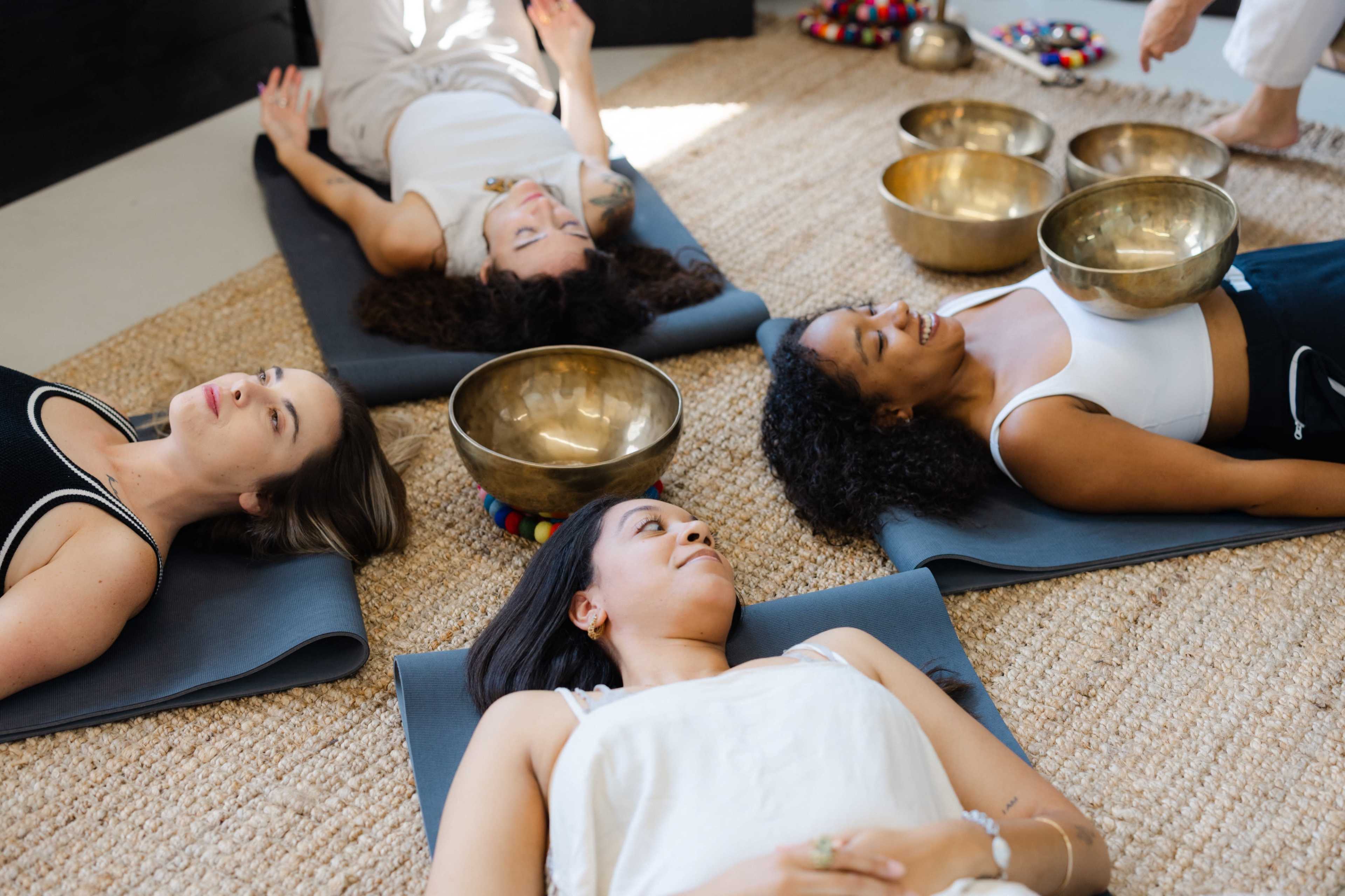 Four women lie on yoga mats in a serene indoor setting, while metal singing bowls are placed nearby.