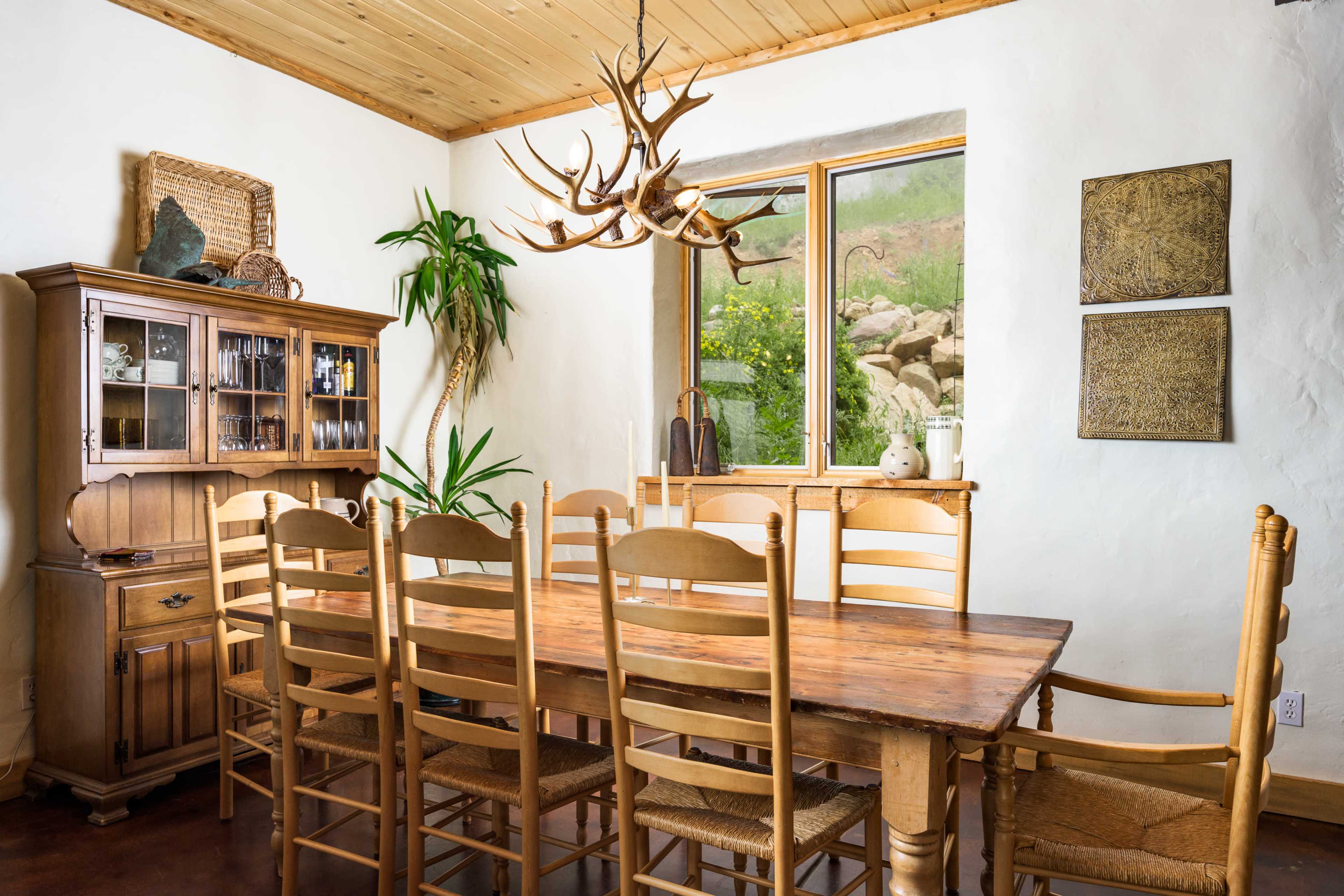 A spacious dining room features a wooden table surrounded by chairs, with an antler chandelier and a large window overlooking greenery.