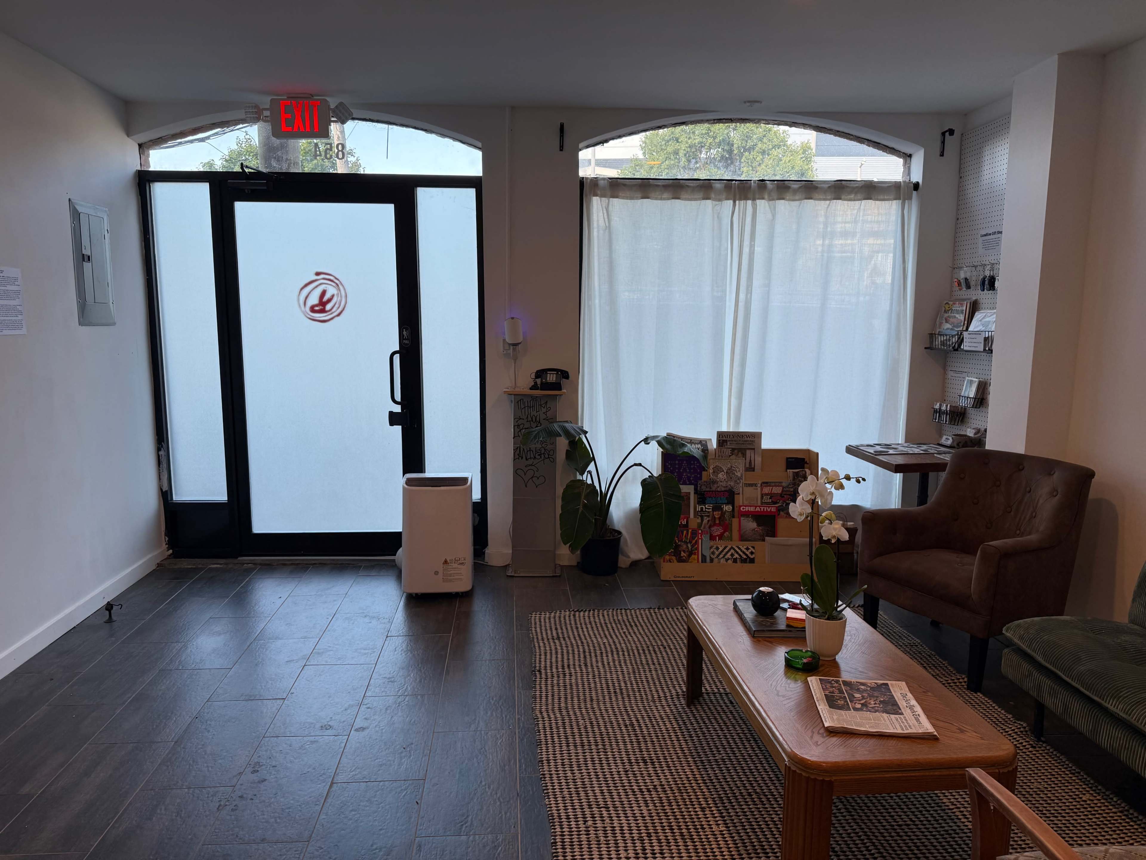 The image shows a modern reception area featuring a door with a circular logo, a seating area with an armchair, a small table, and various plants and reading materials.