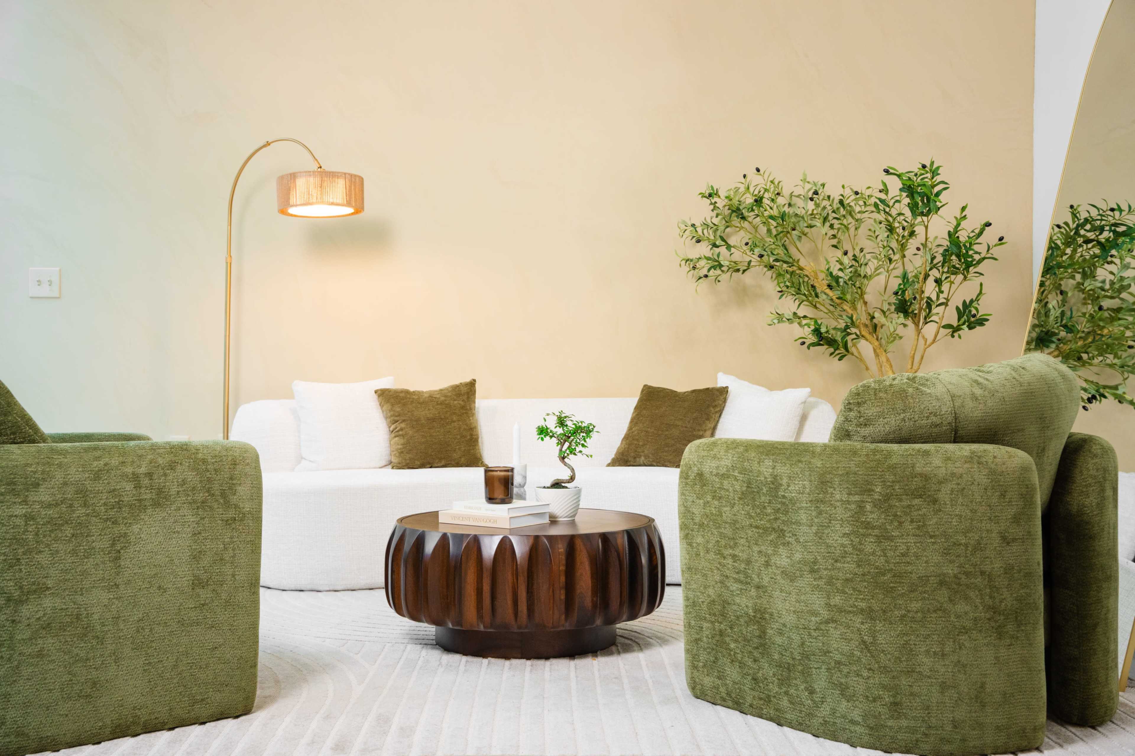 The image shows a modern living room with a white sectional sofa, two green armchairs, a circular wooden coffee table, and a floor lamp, all set against a light-colored wall.