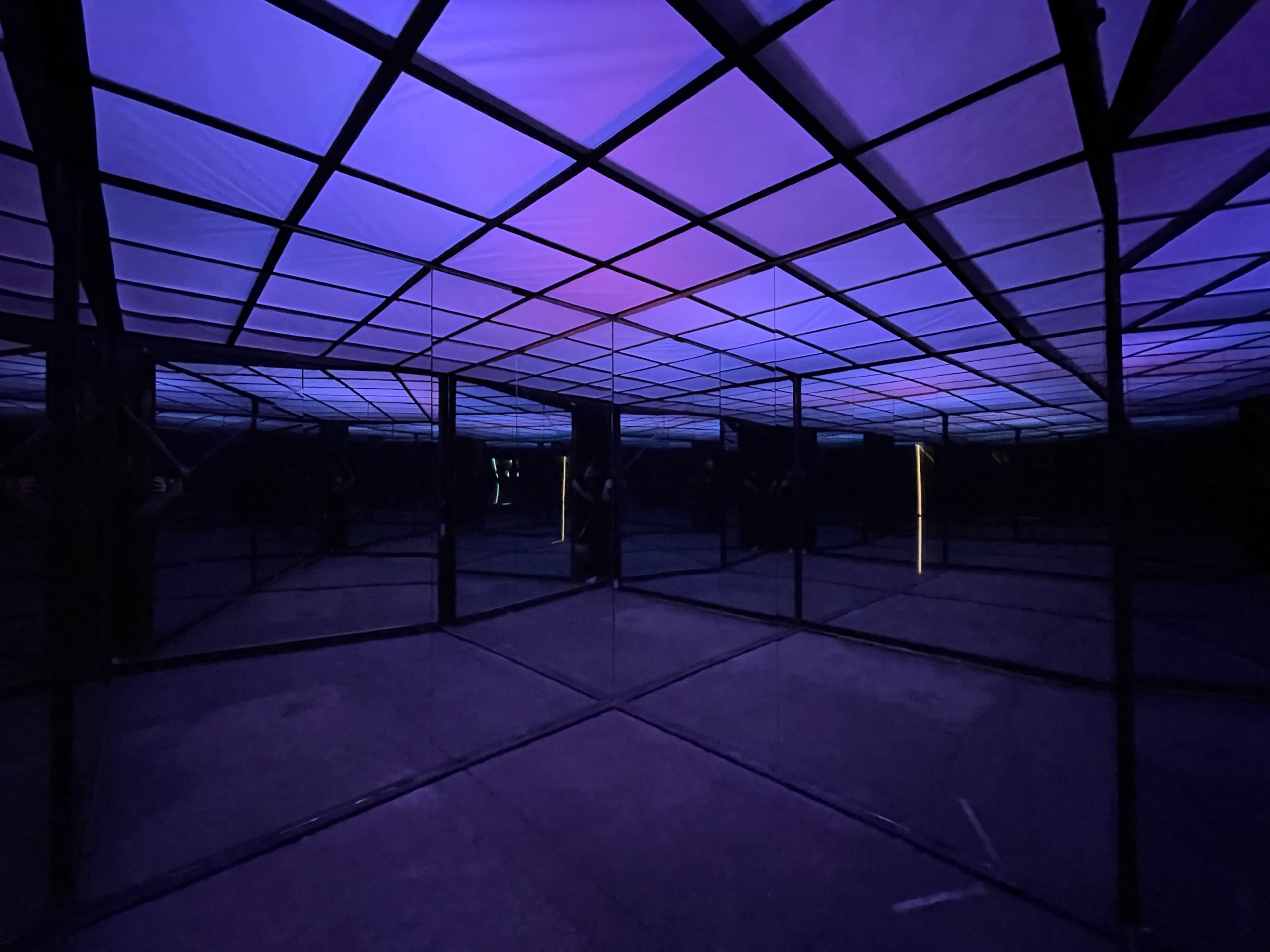 A geometric mirror room features a grid-patterned ceiling illuminated in purple and blue hues.