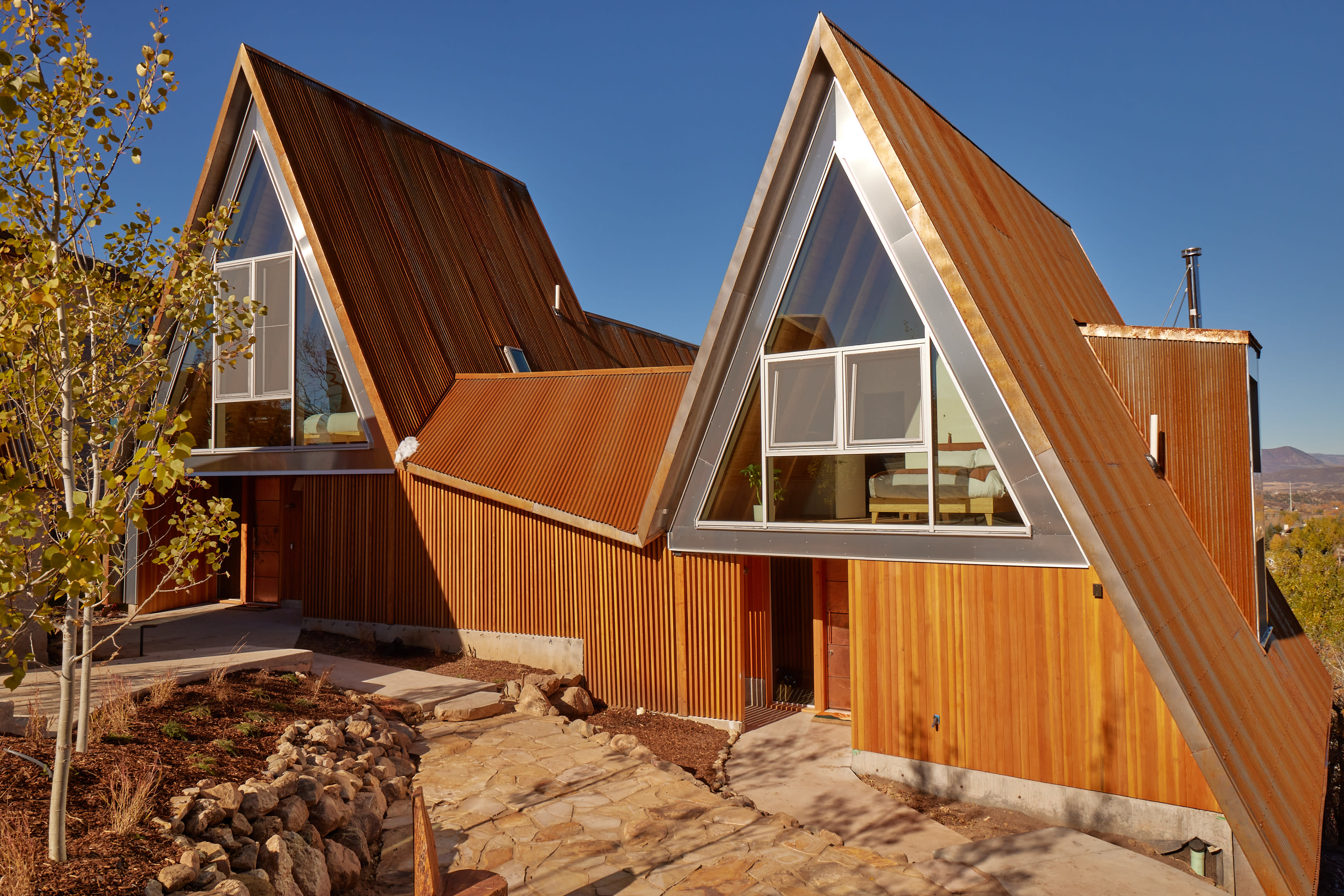 A contemporary A-frame house with a wooden exterior and large triangular windows is set against a clear blue sky.