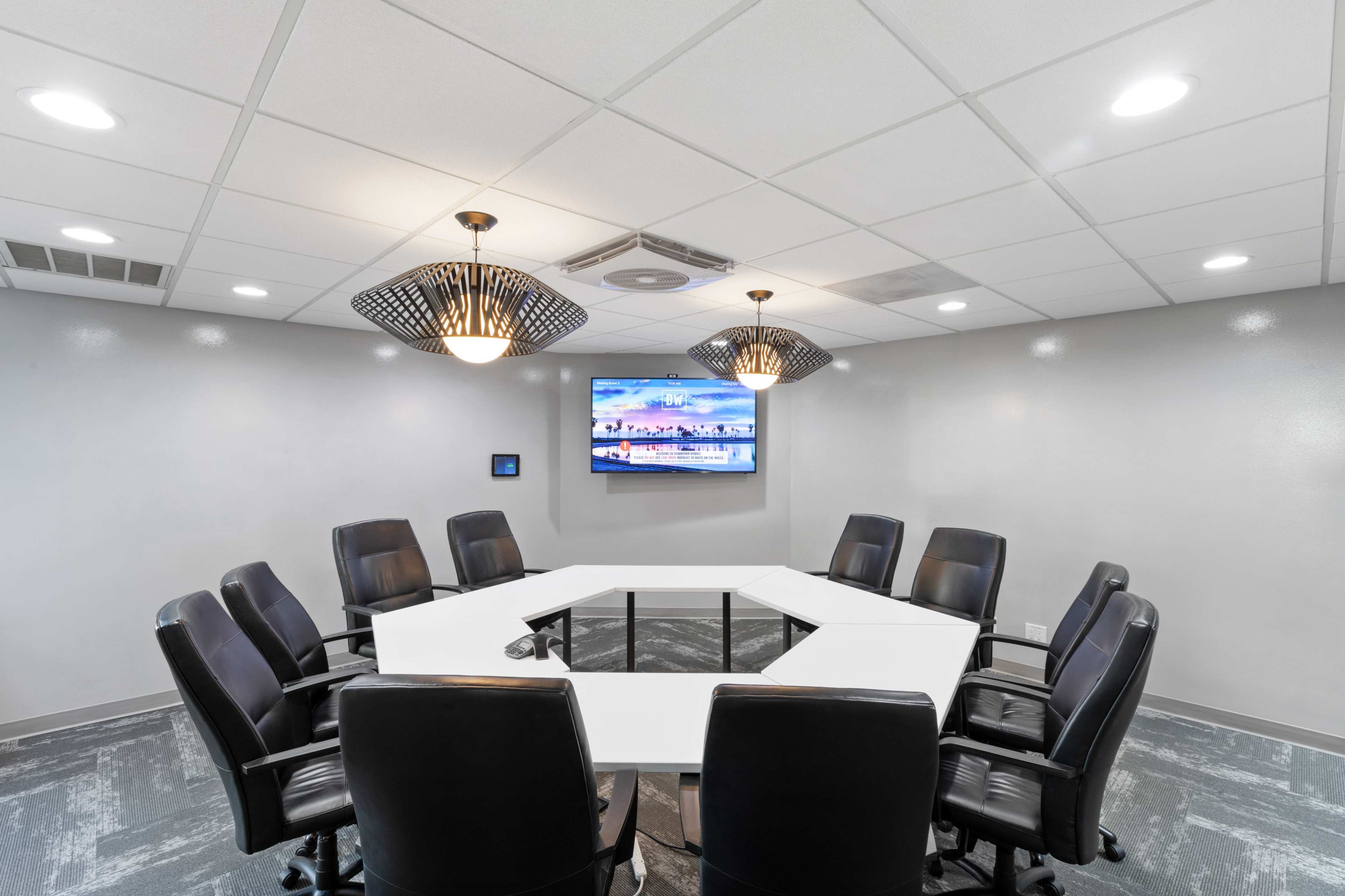 A modern conference room features a large, white octagonal table surrounded by black leather chairs, with a flat-screen TV mounted on the wall.