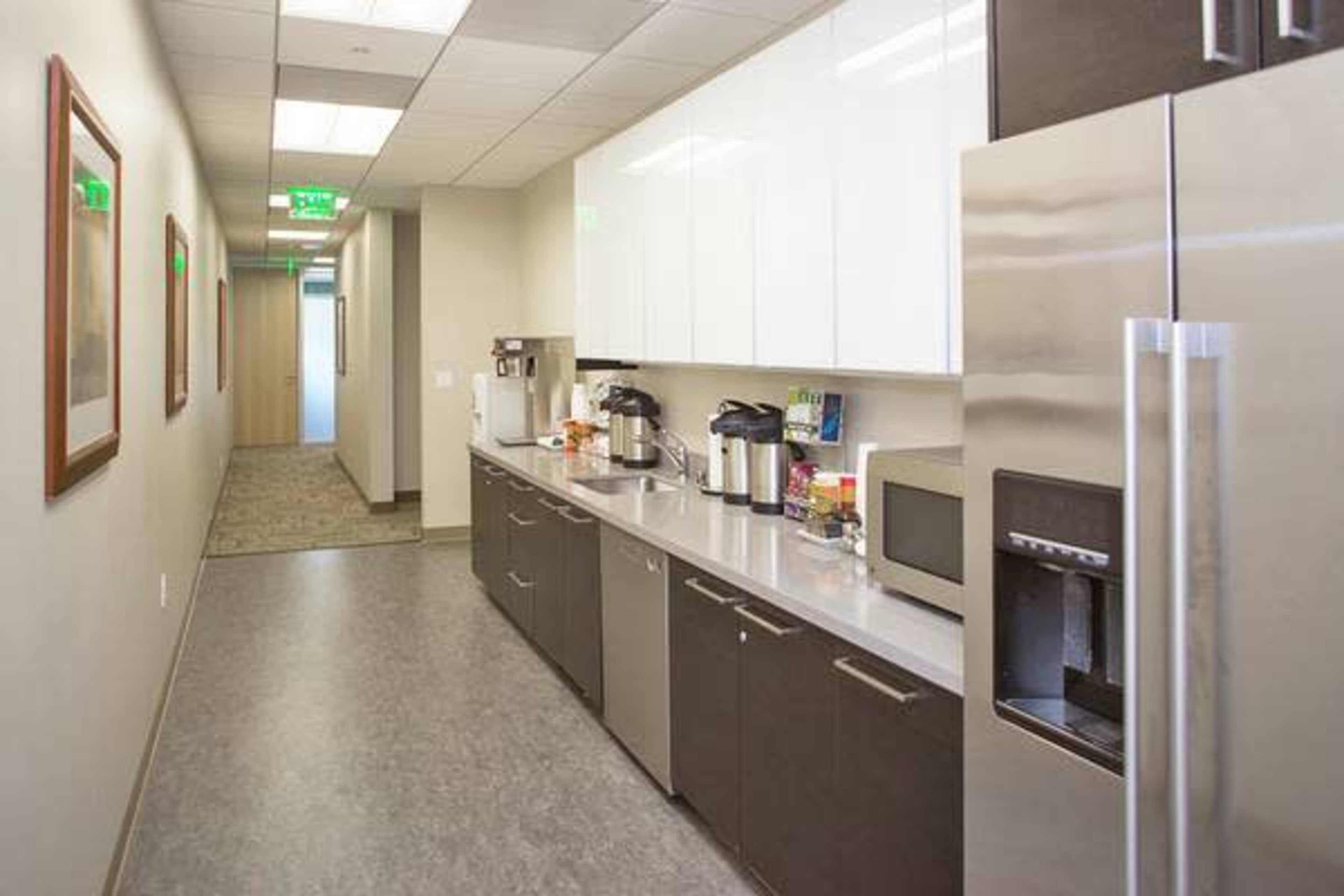 A modern office kitchenette with a long counter, coffee machines, a microwave, and stainless steel appliances along a corridor.
