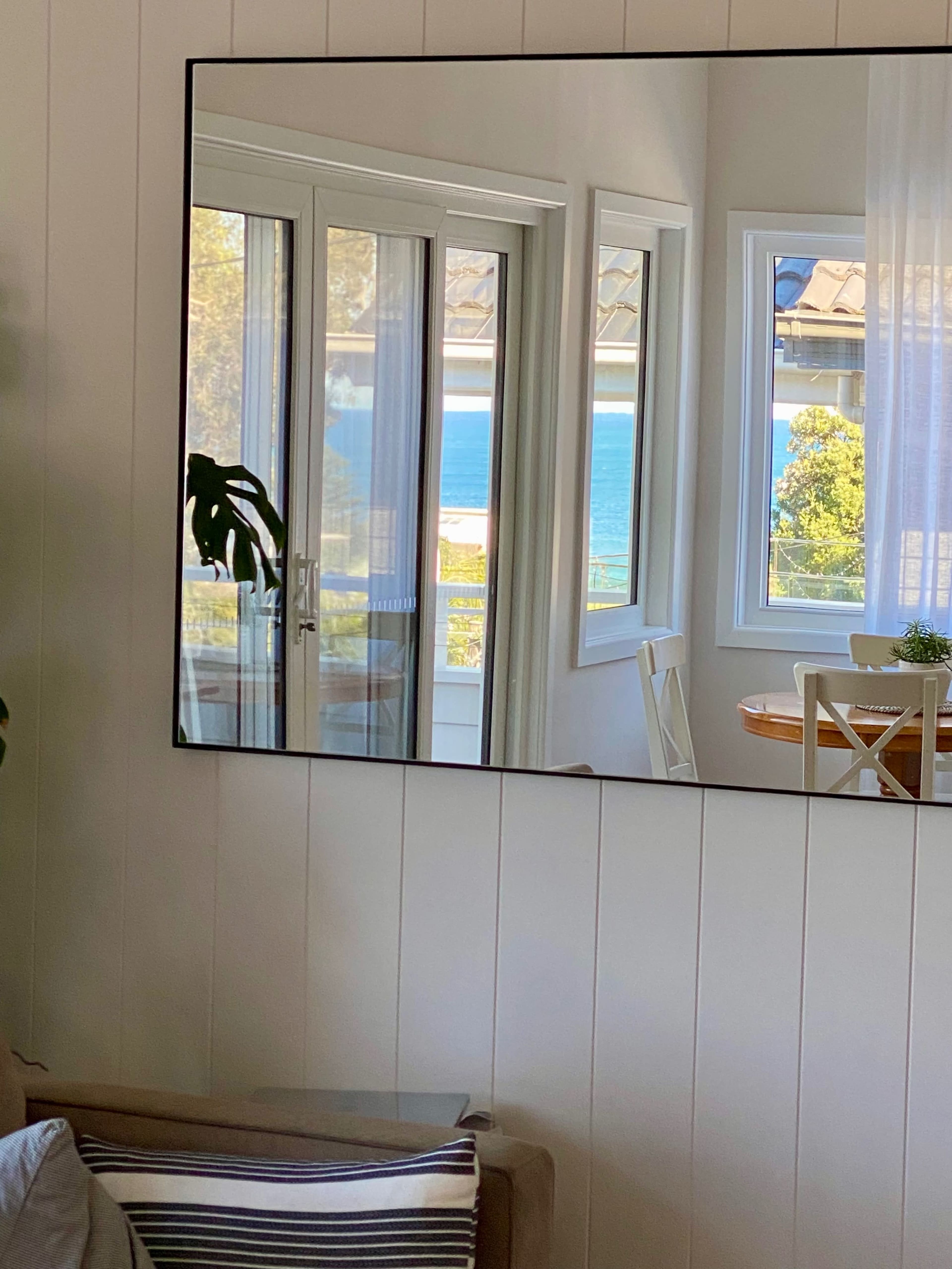 A large mirror reflects a view of the ocean visible through a set of windows in a bright room.