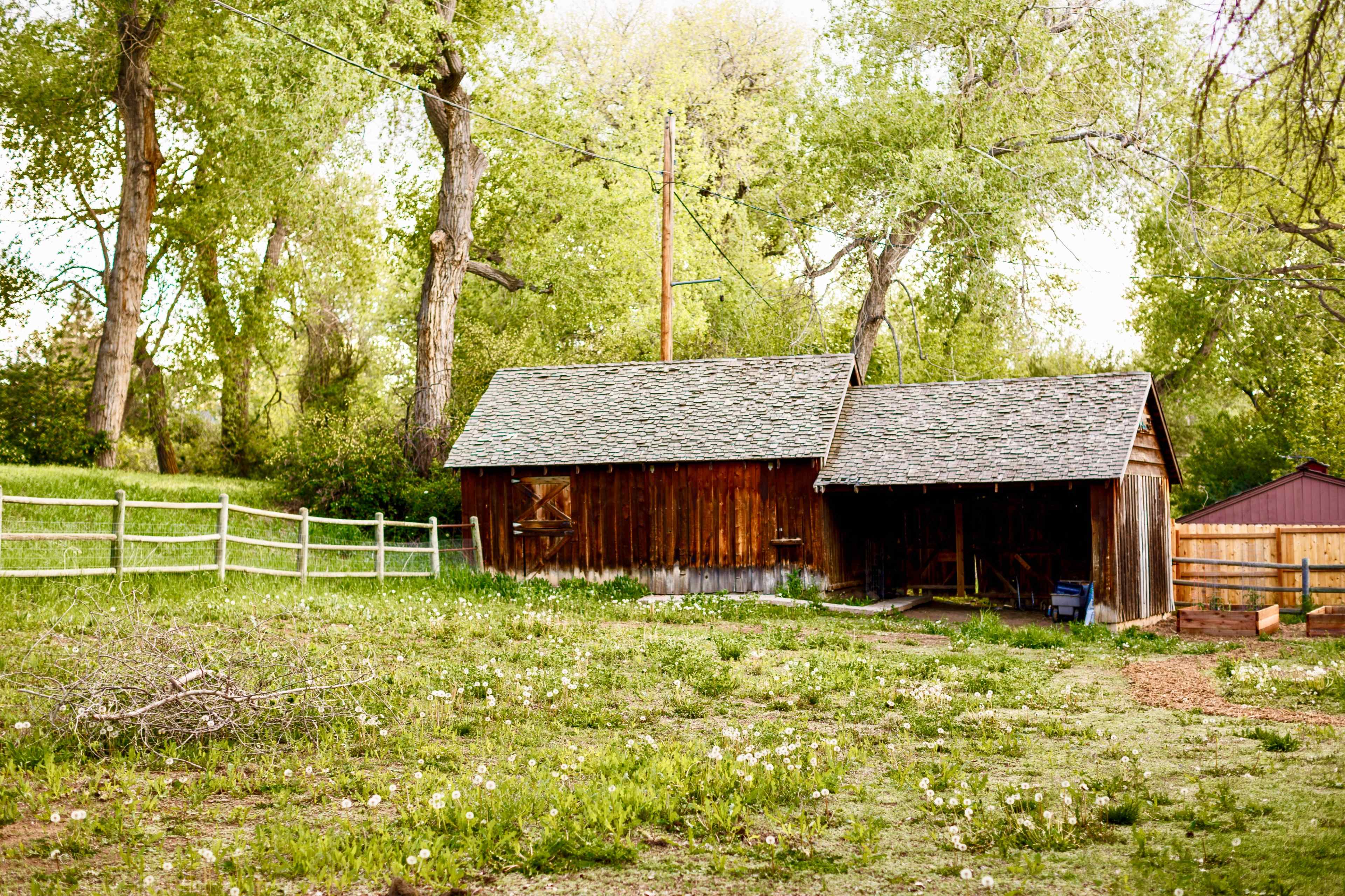 A wooden barn sits on a grassy area surrounded by trees and a fence.