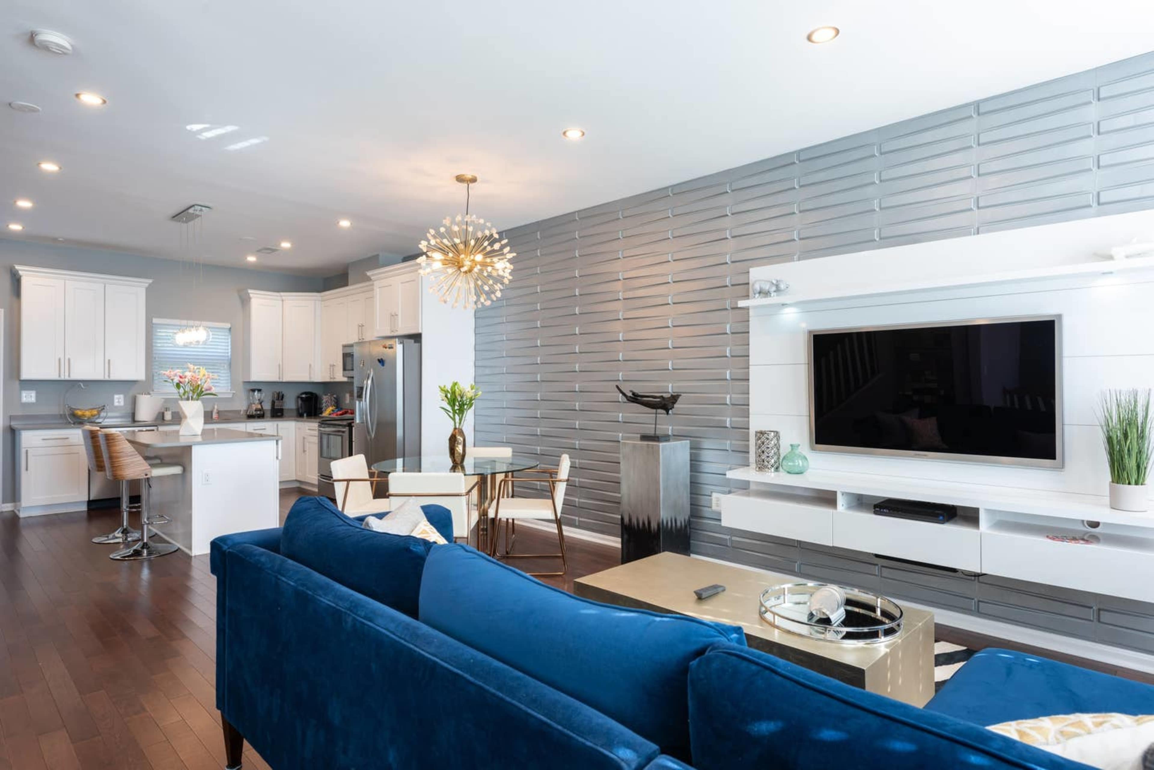 A modern living room featuring a blue sofa, a dining area with a round table, and a flat-screen television mounted on a textured wall.
