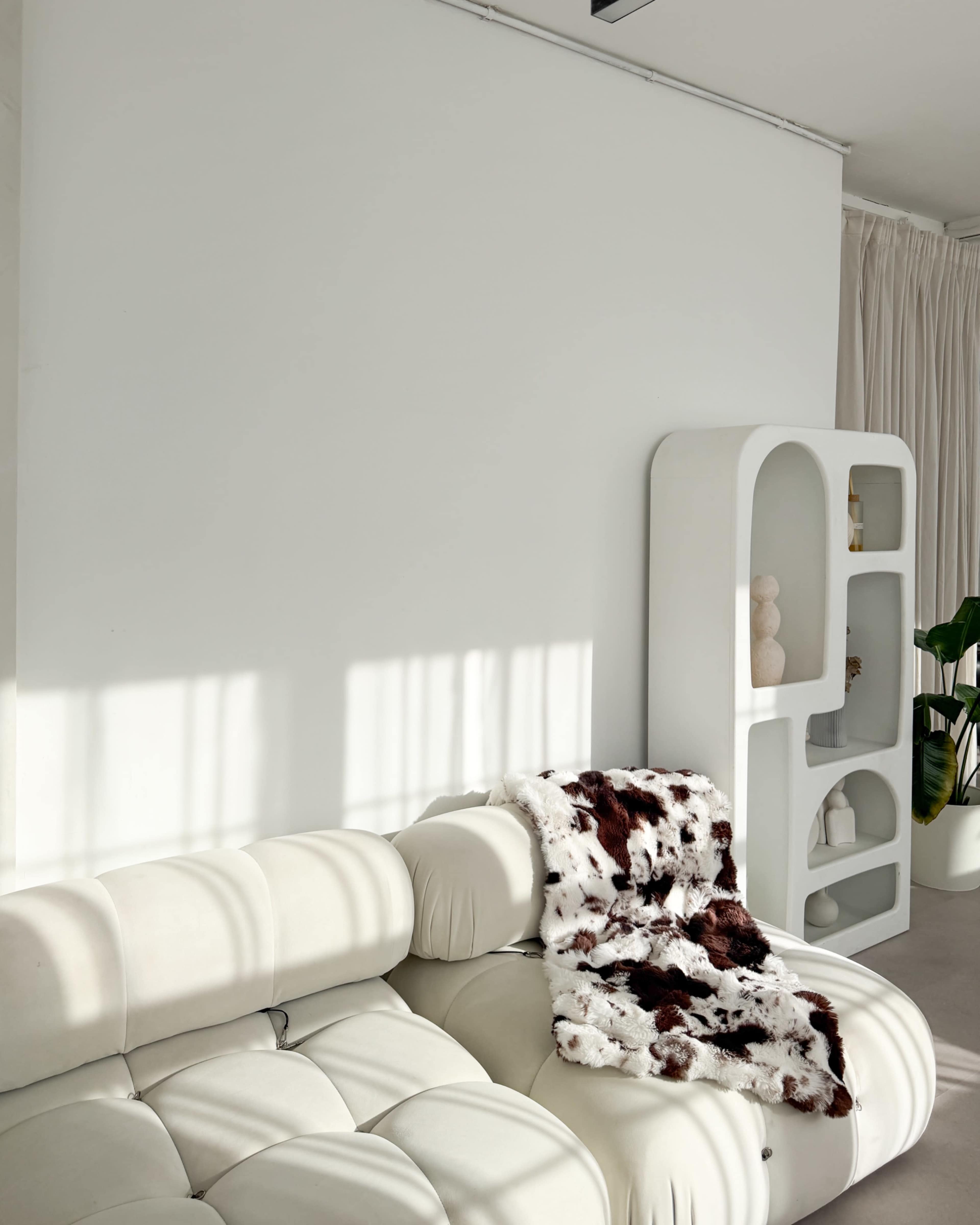 The image shows a white sofa with a patterned throw draped over it, alongside a modern white shelving unit in a well-lit interior space.