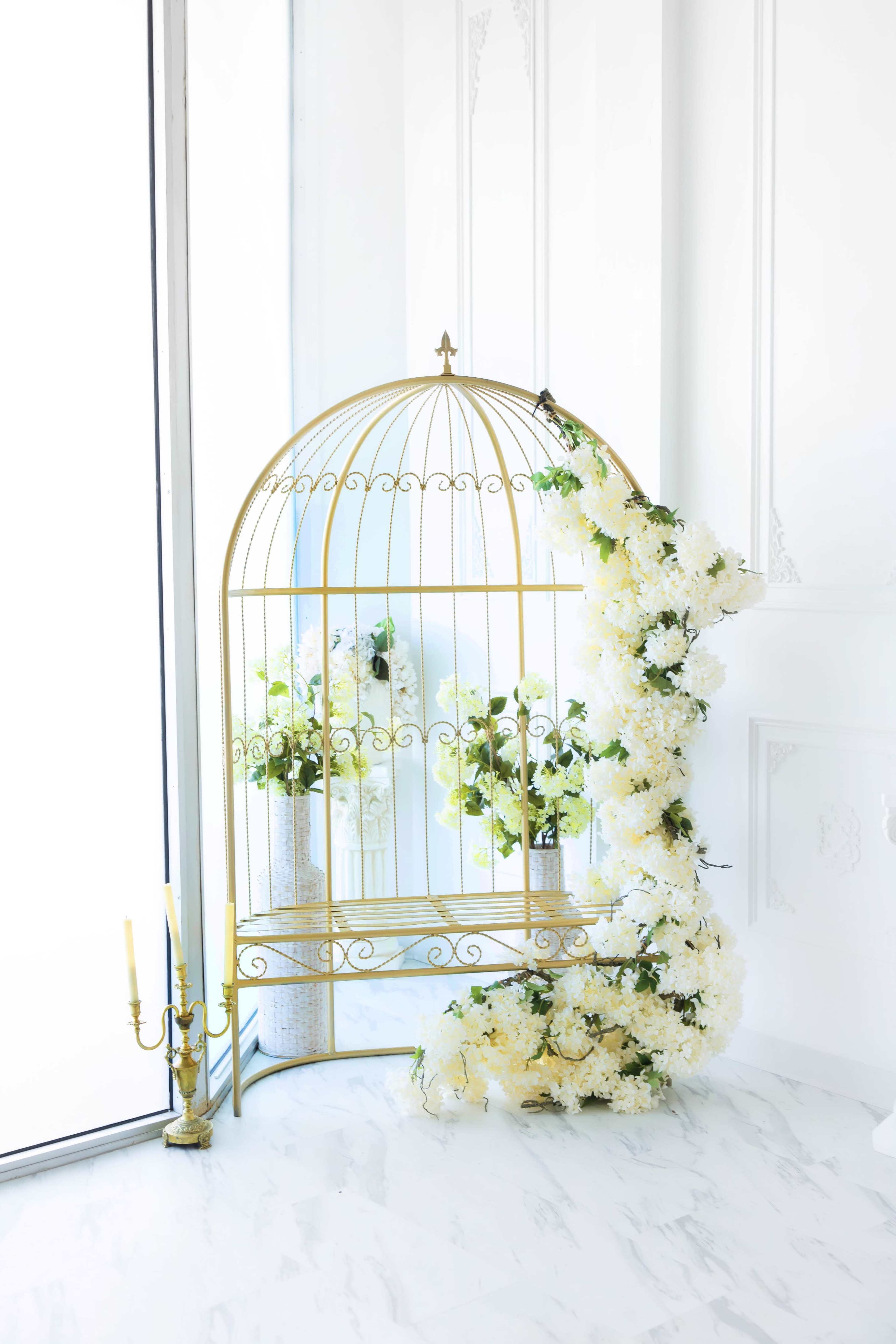 A gold metal birdcage stands beside a tall vase filled with greenery and white flowers, with additional floral arrangements cascading down its side.