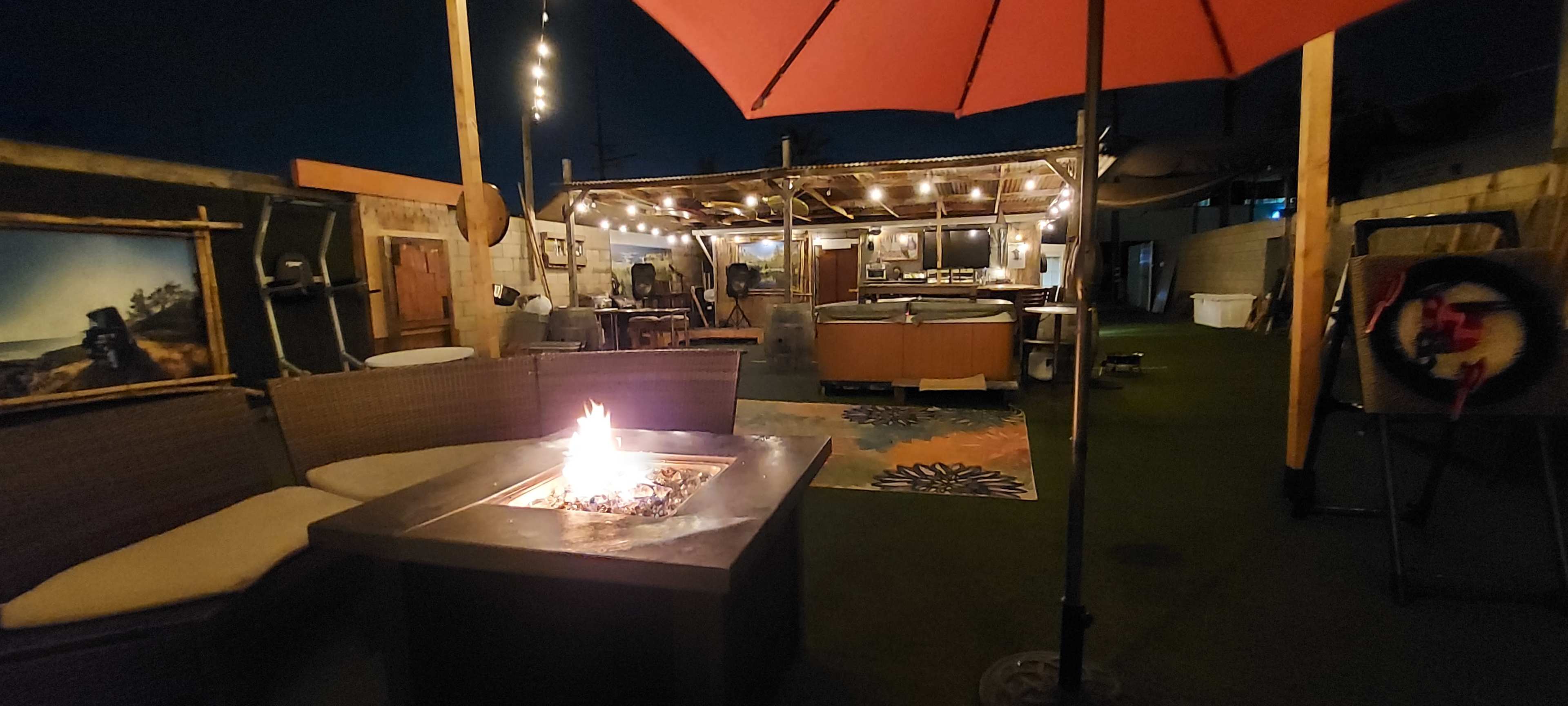 A cozy outdoor patio area with a fire pit, seating around it, and a well-lit kitchen area in the background under a canopy.