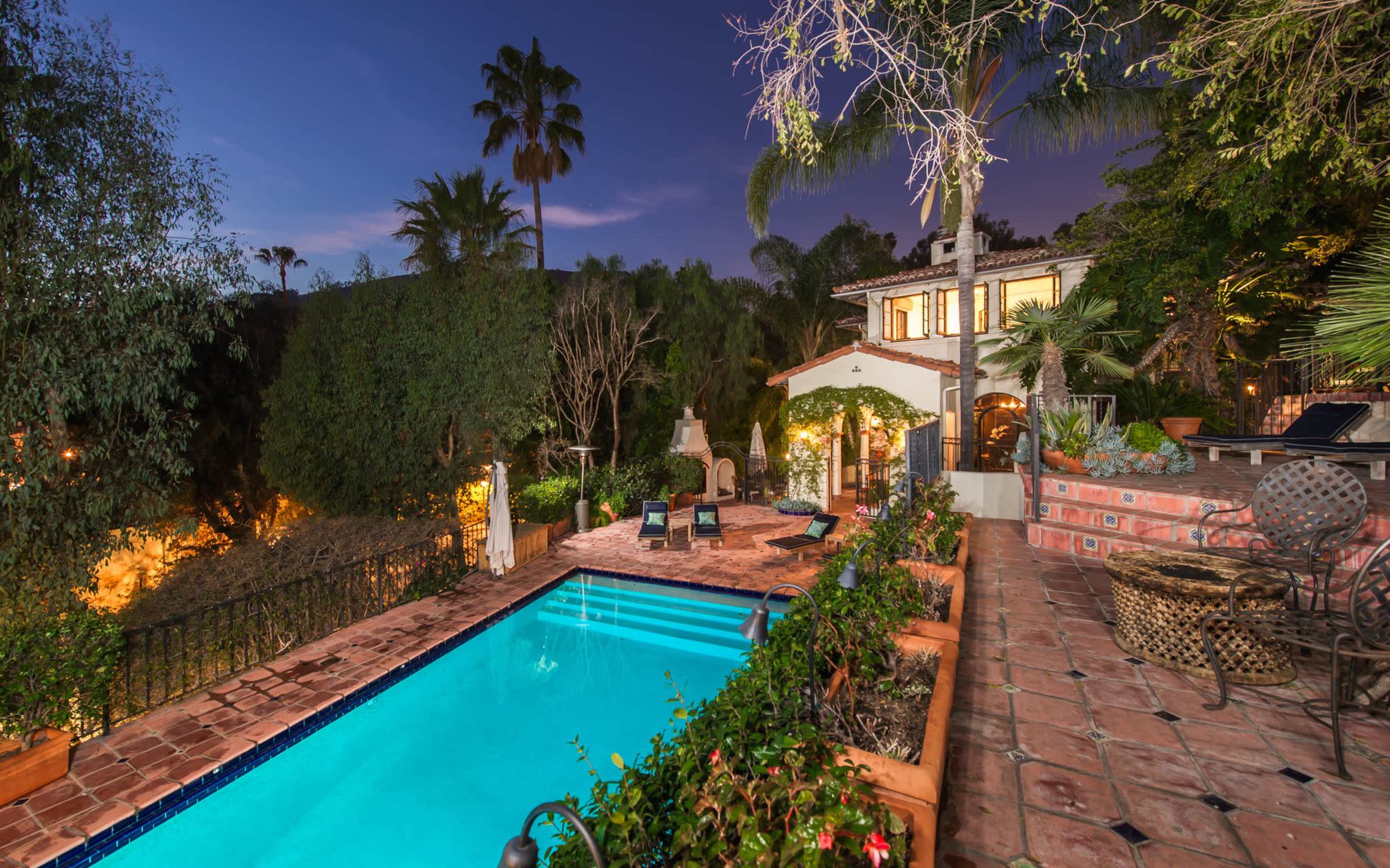 A Spanish-style villa with a swimming pool is surrounded by lush greenery and palm trees at dusk.