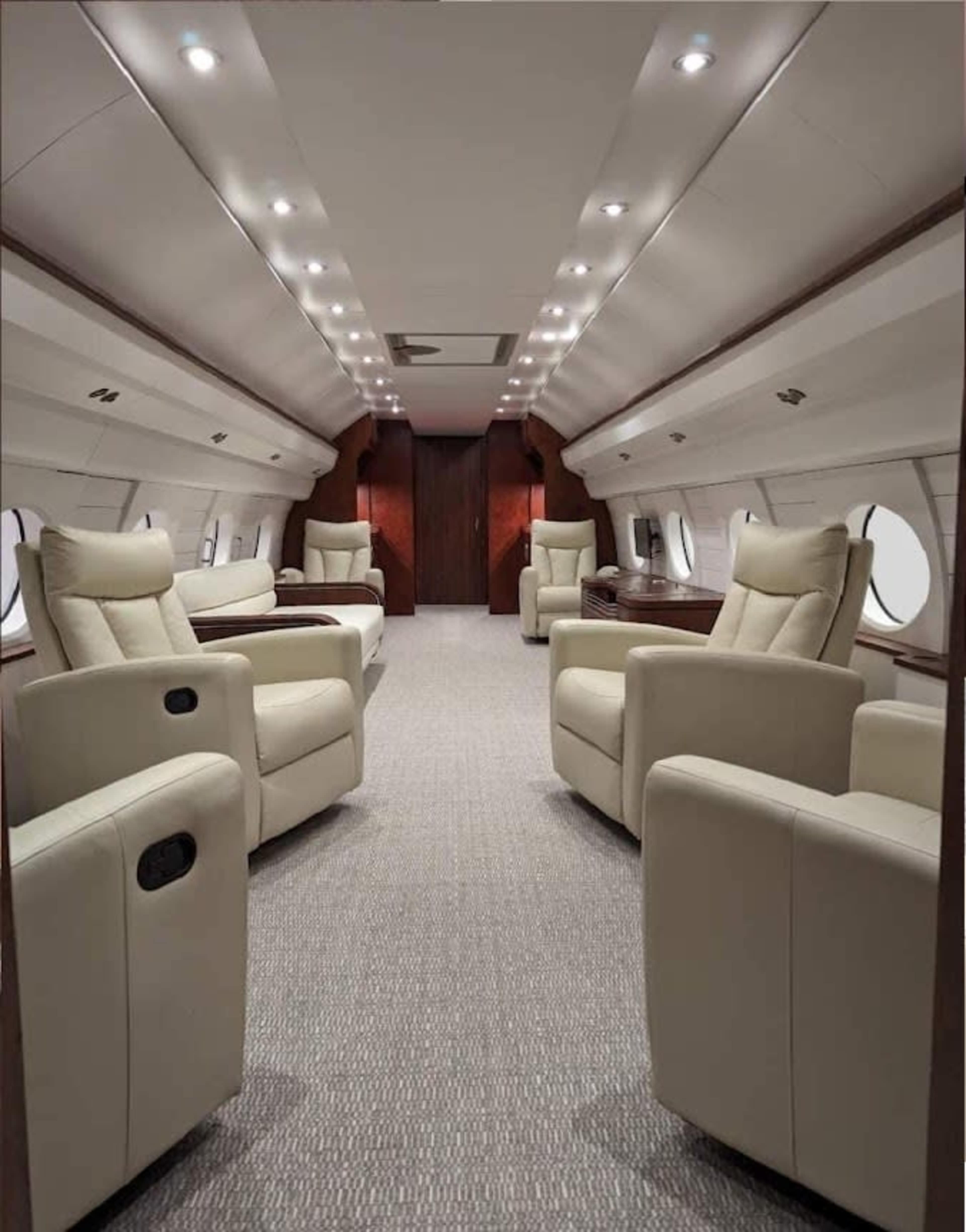 The interior of a private jet features plush beige reclining chairs arranged along the sides with large windows and a sleek, polished wooden door at the far end.