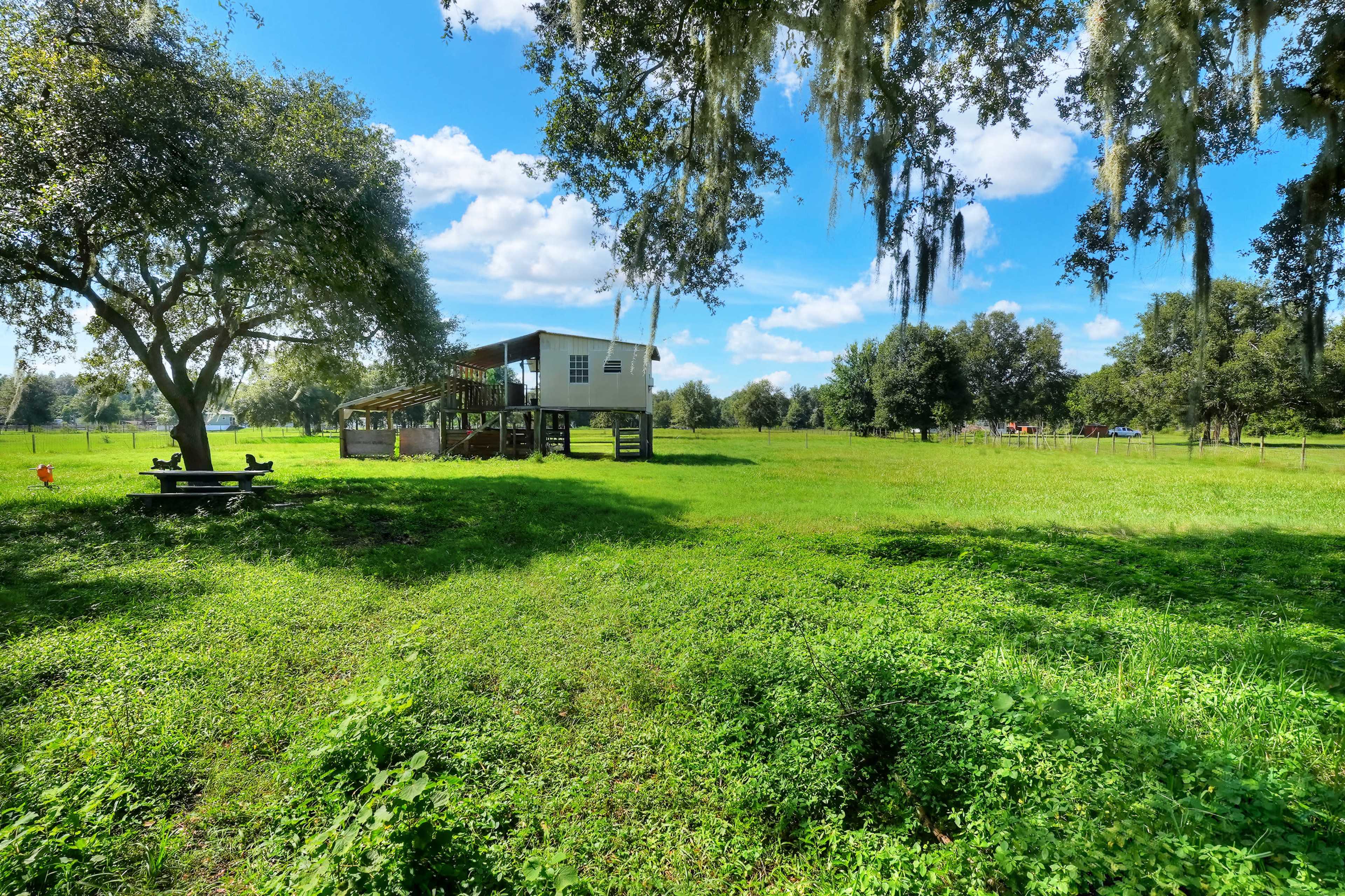 A small elevated house sits on stilts in a green field surrounded by trees and under a clear blue sky.