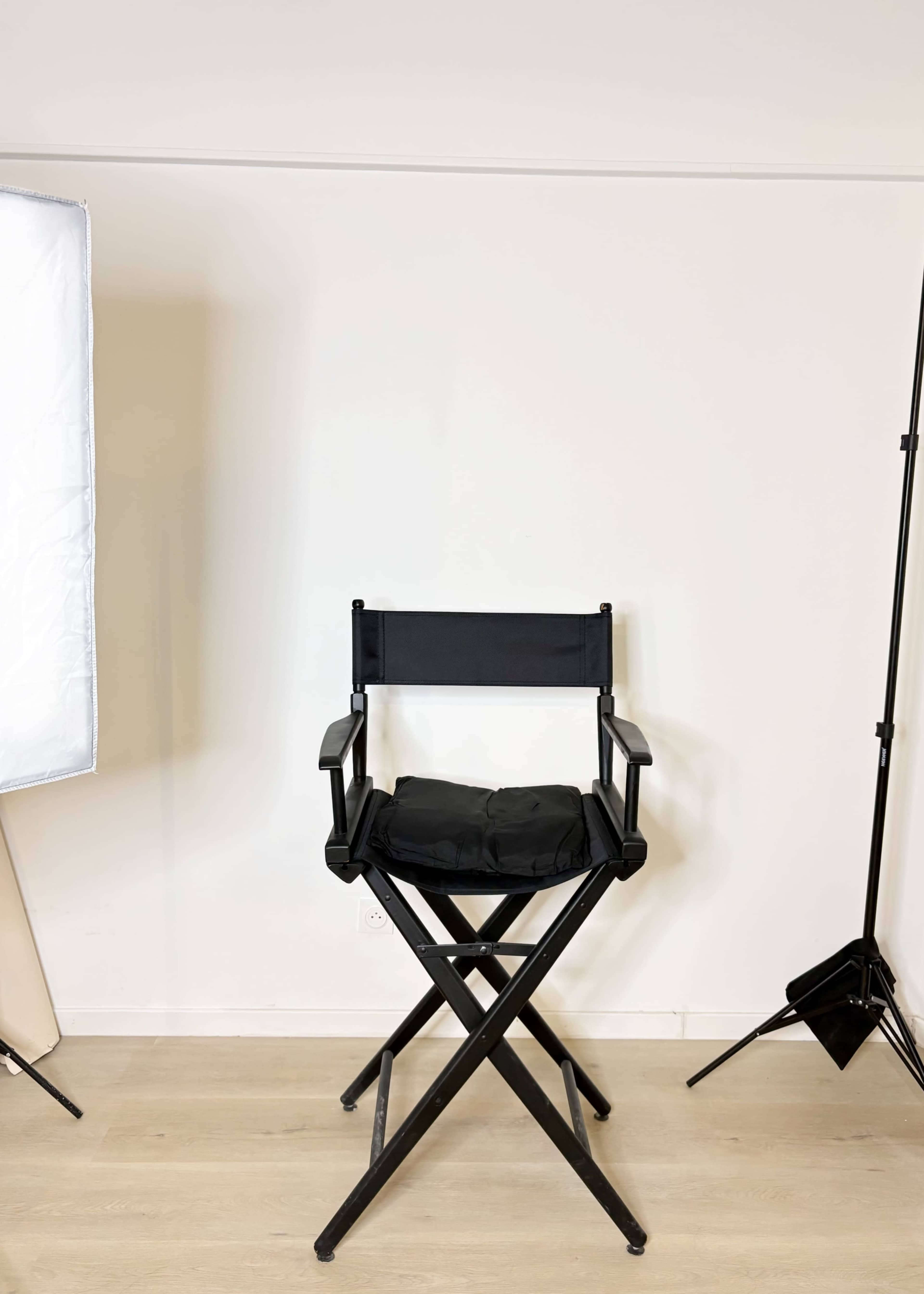 The image shows a black director's chair positioned against a plain white wall, with studio lighting equipment nearby.