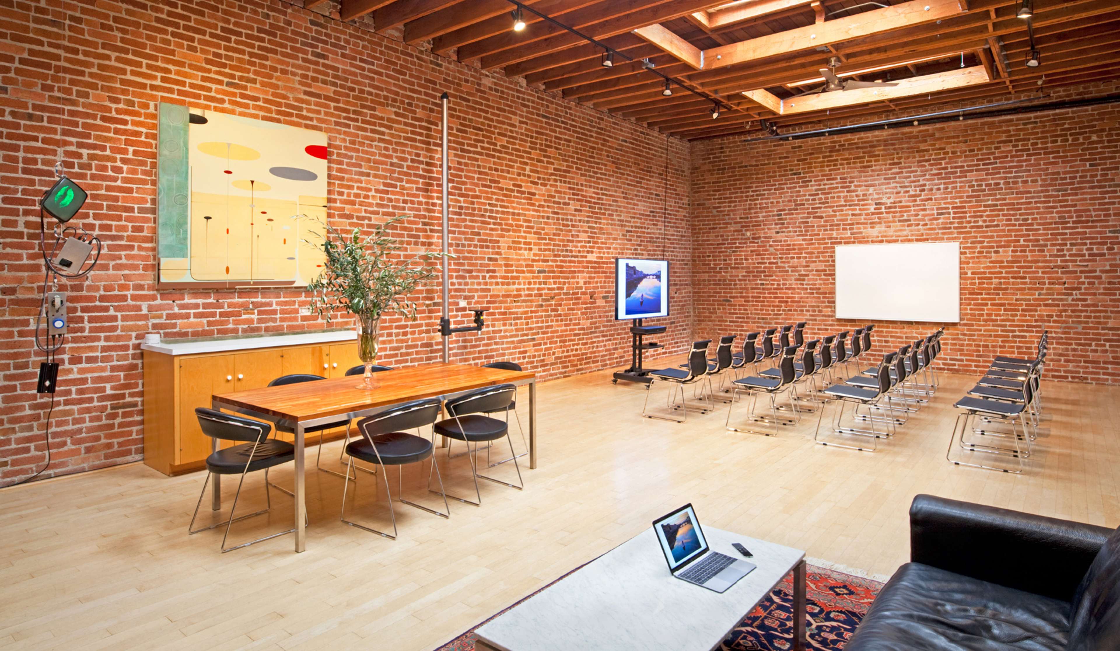 A spacious meeting room with a wooden table and chairs, exposed brick walls, and a screen at the front, along with a couch and a coffee table.
