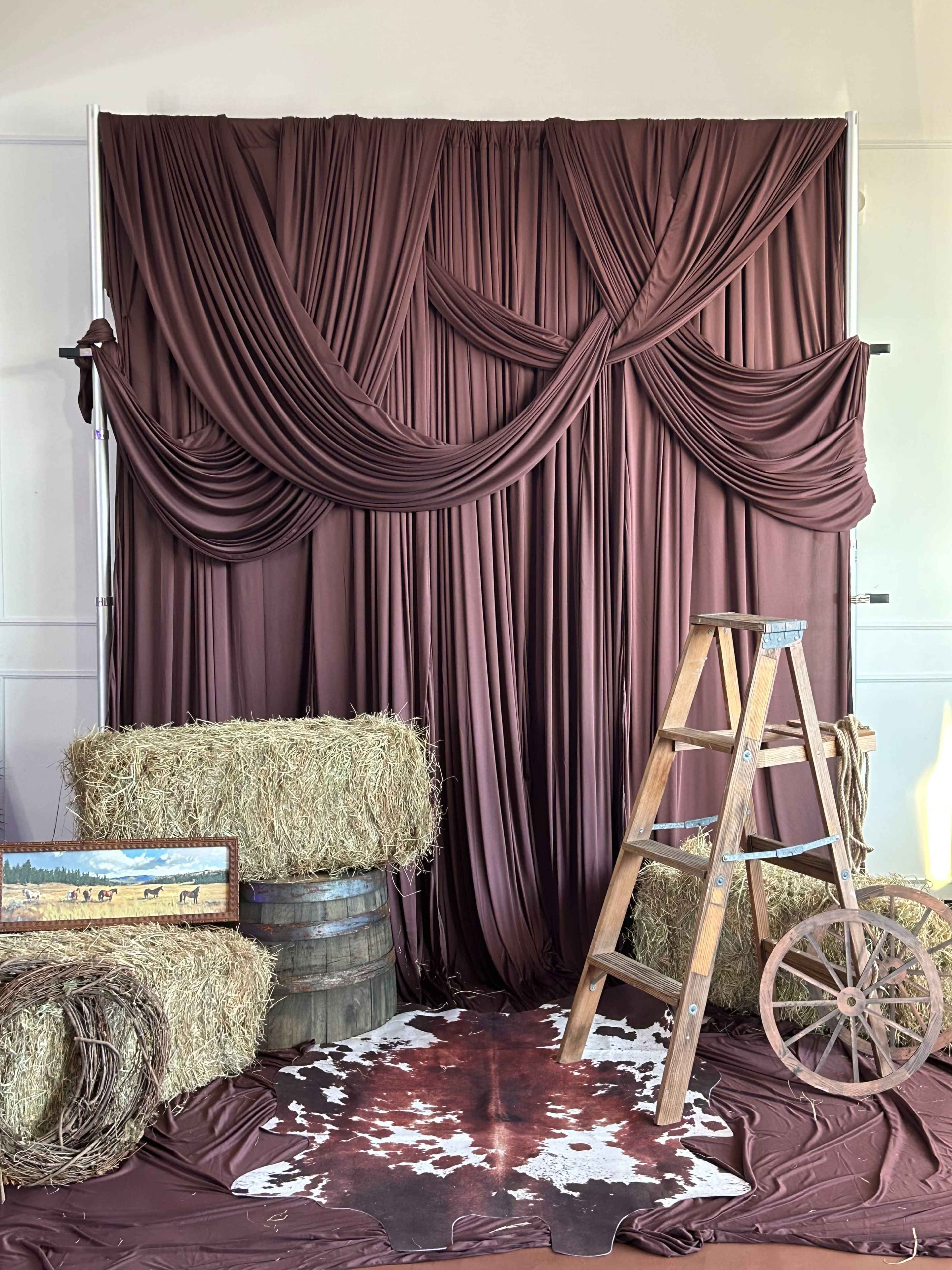 The image features a rustic setting with a brown draped backdrop, bales of hay, a wooden ladder, a barrel, and a cowhide rug.