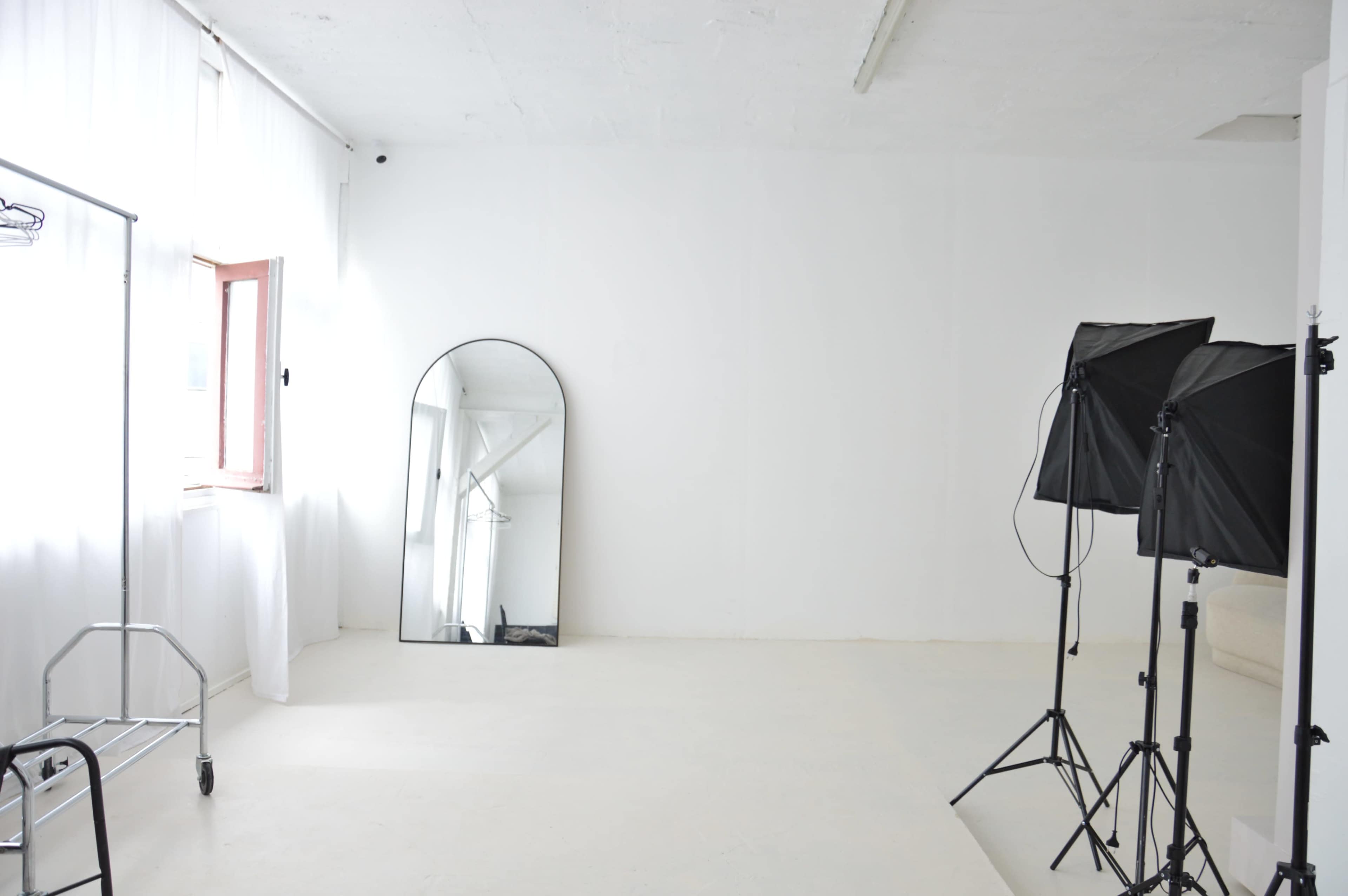 The image shows a bright, empty room with white walls and floor, featuring a full-length mirror and a light setup on a tripod.