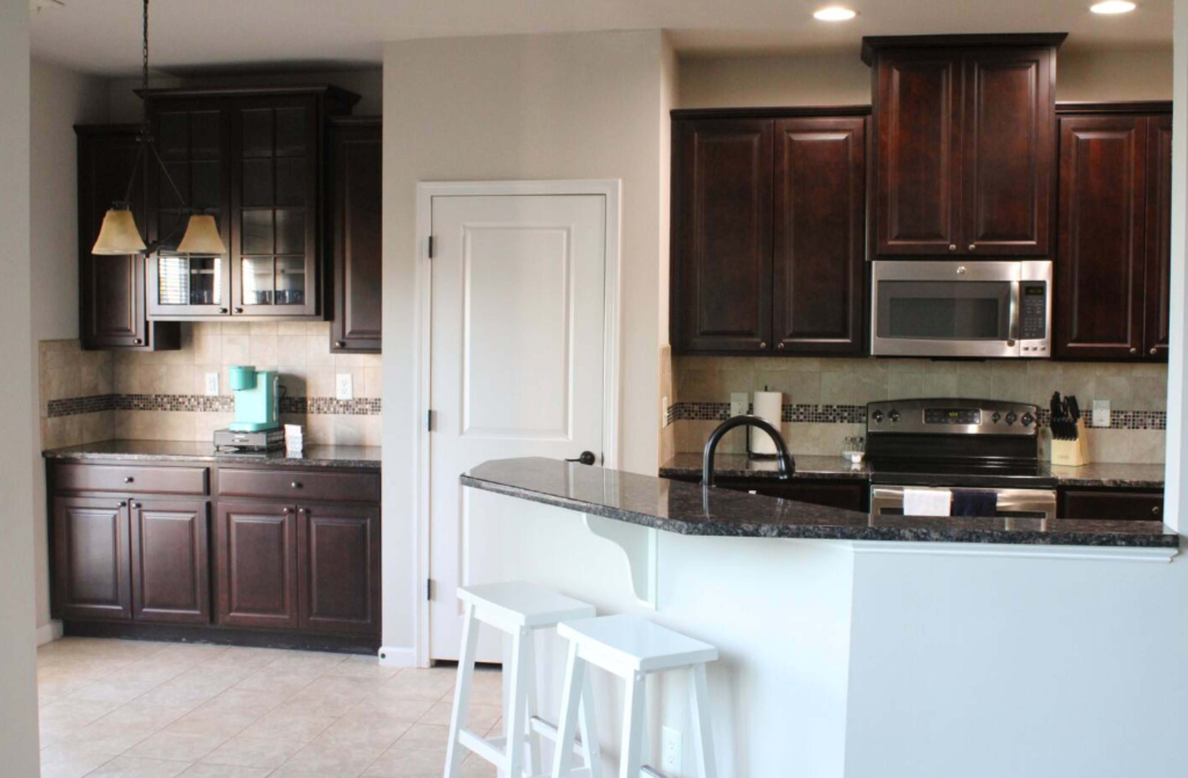 The image shows a modern kitchen with dark wood cabinets, stainless steel appliances, and a granite countertop with bar stools.