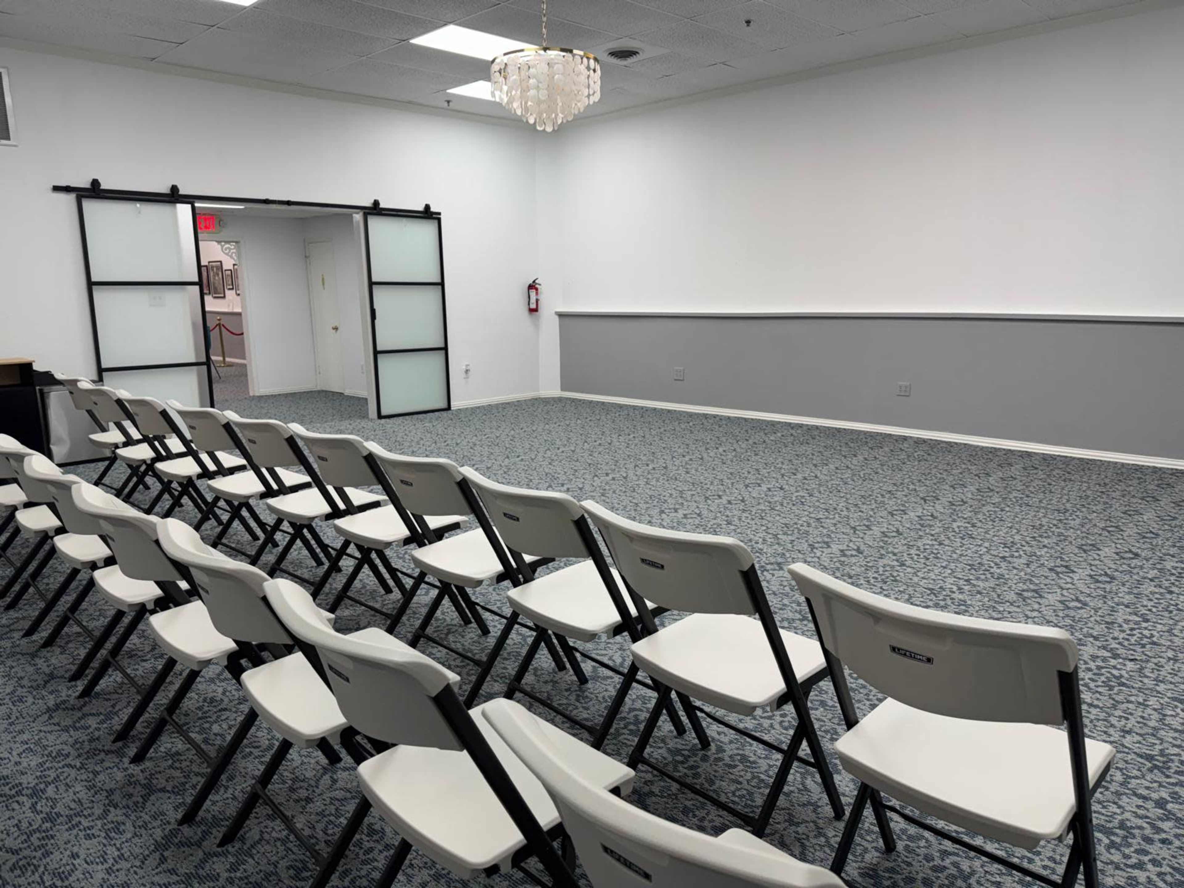 A row of white folding chairs faces a blank wall in a spacious room with a patterned carpet and a chandelier overhead.