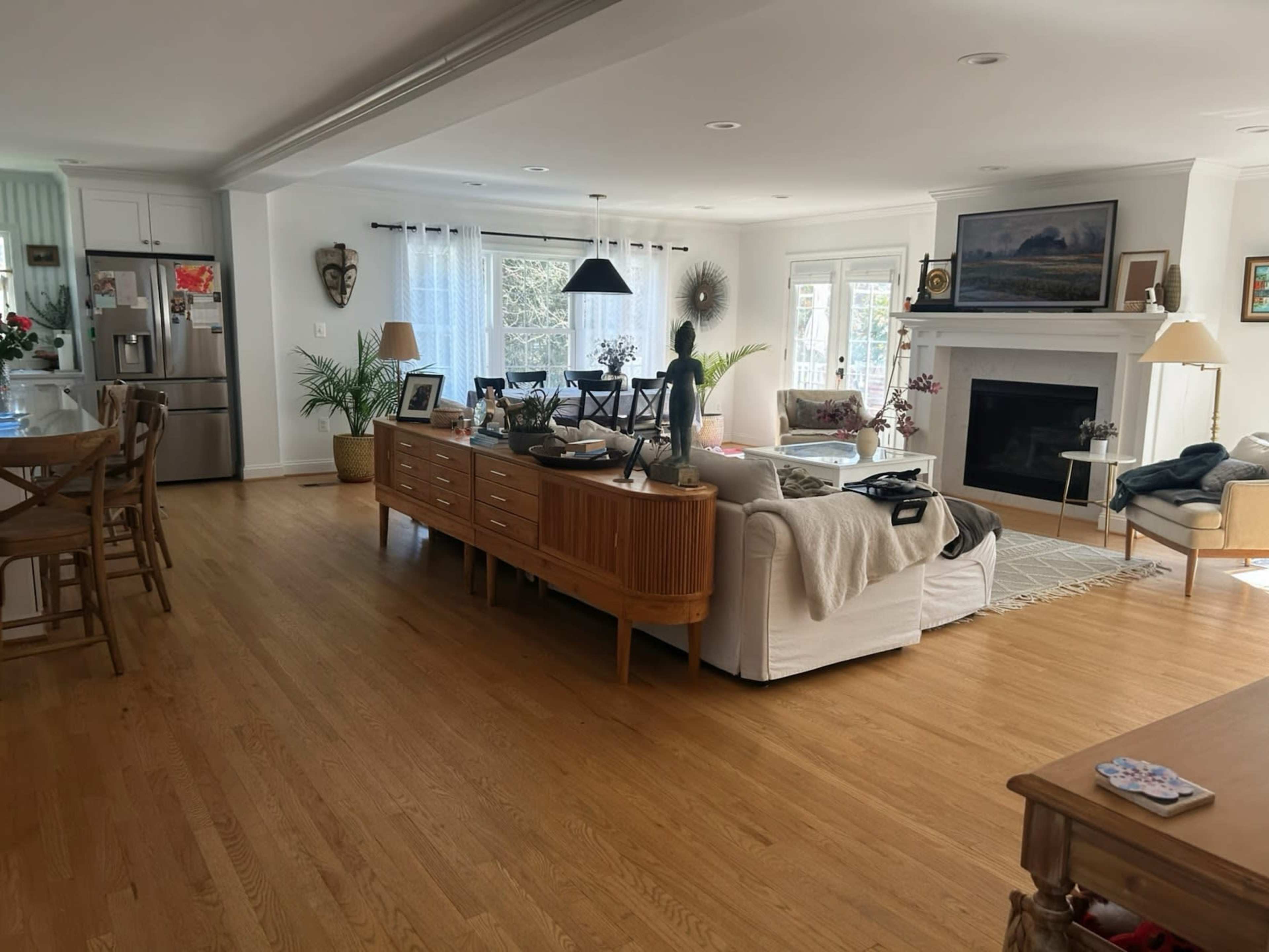 The image shows a bright, spacious living room with hardwood floors, a large window, and a mix of furniture including a sectional sofa, a wooden sideboard, and a cozy armchair.