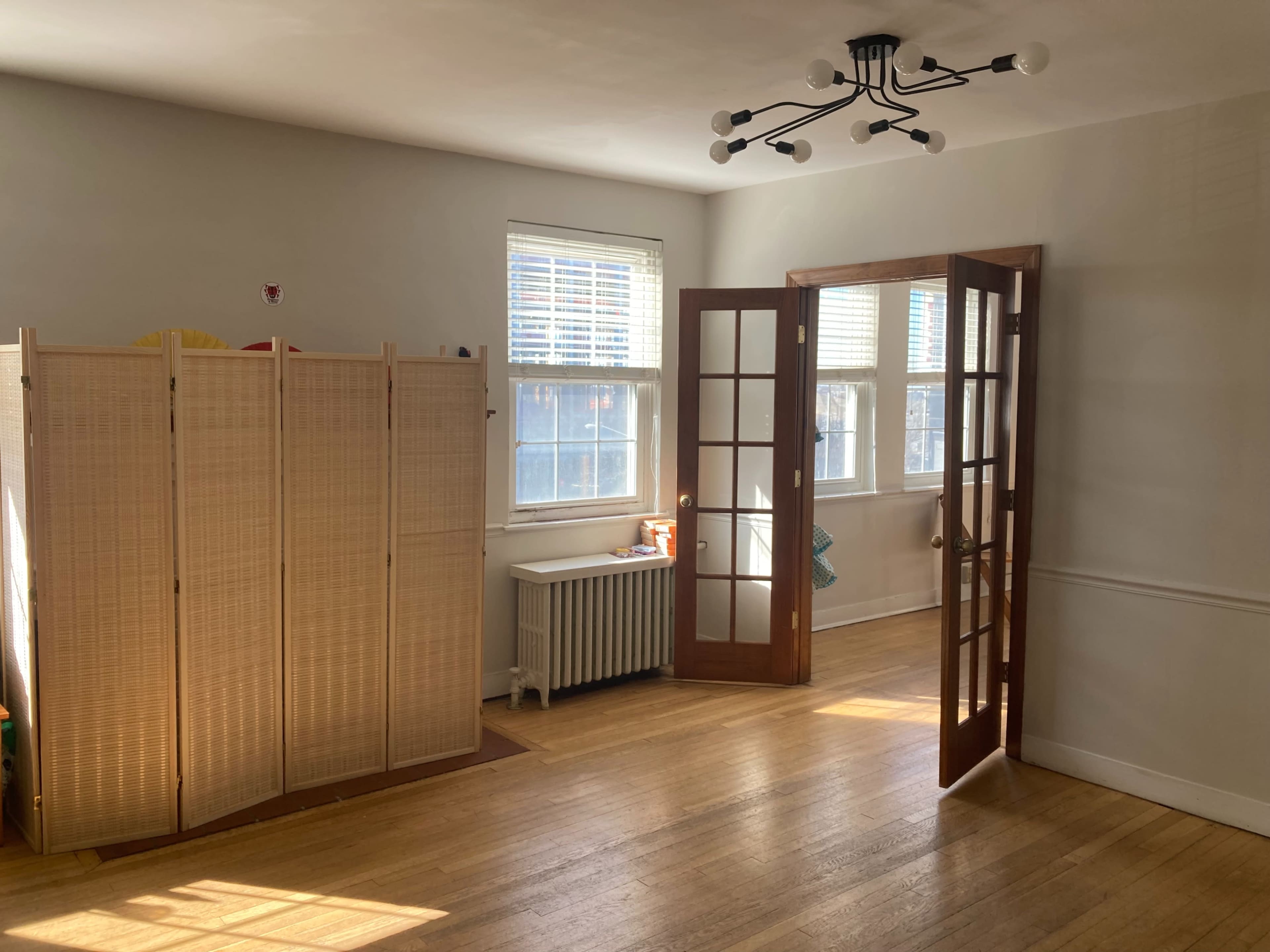 A bright room with wooden floors, featuring a folding screen, a radiator, and double doors open to another space, along with several windows allowing natural light.
