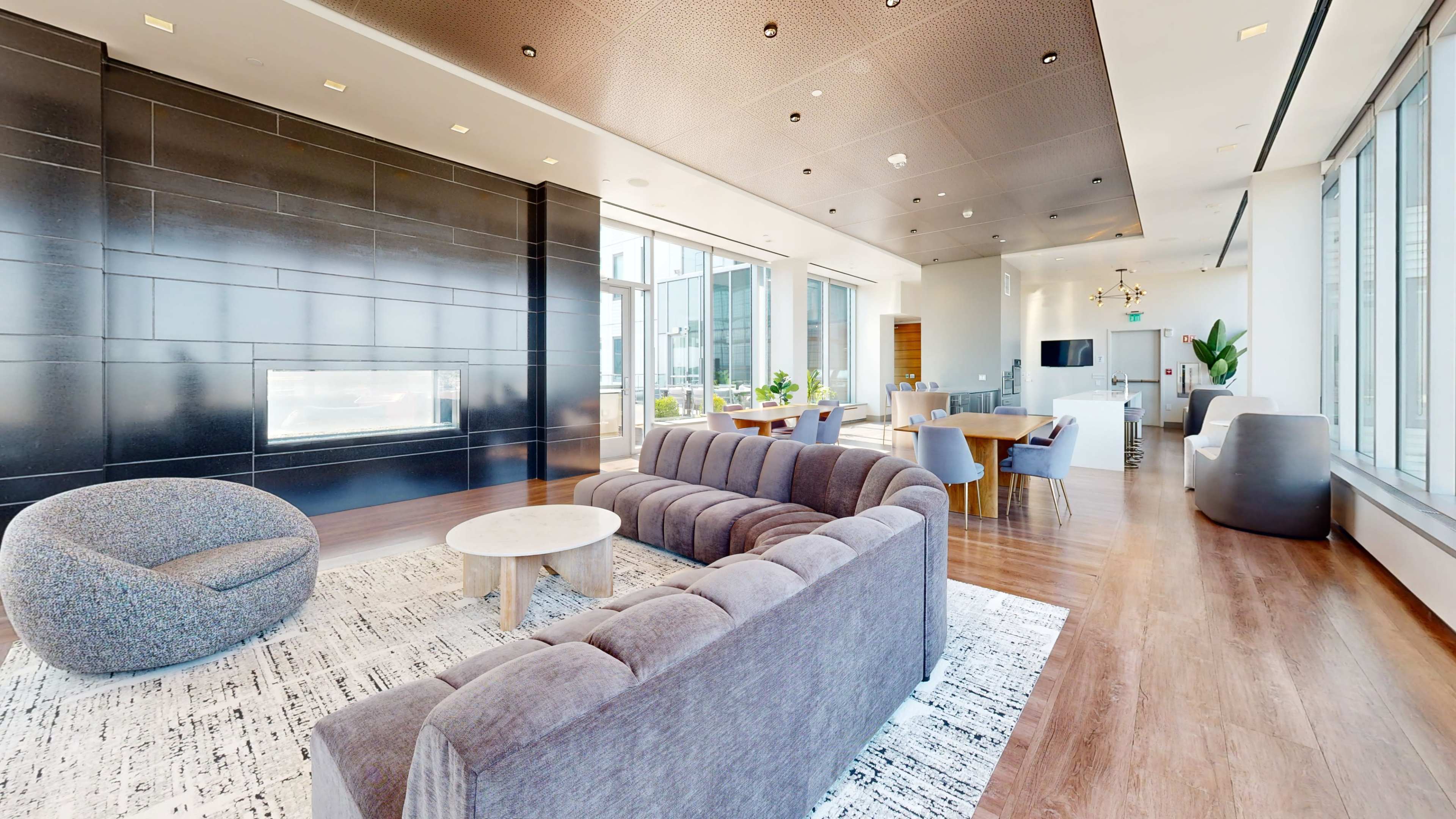 The image shows a modern, open-concept lounge area featuring a gray sectional sofa, a circular coffee table, and large windows with natural light.