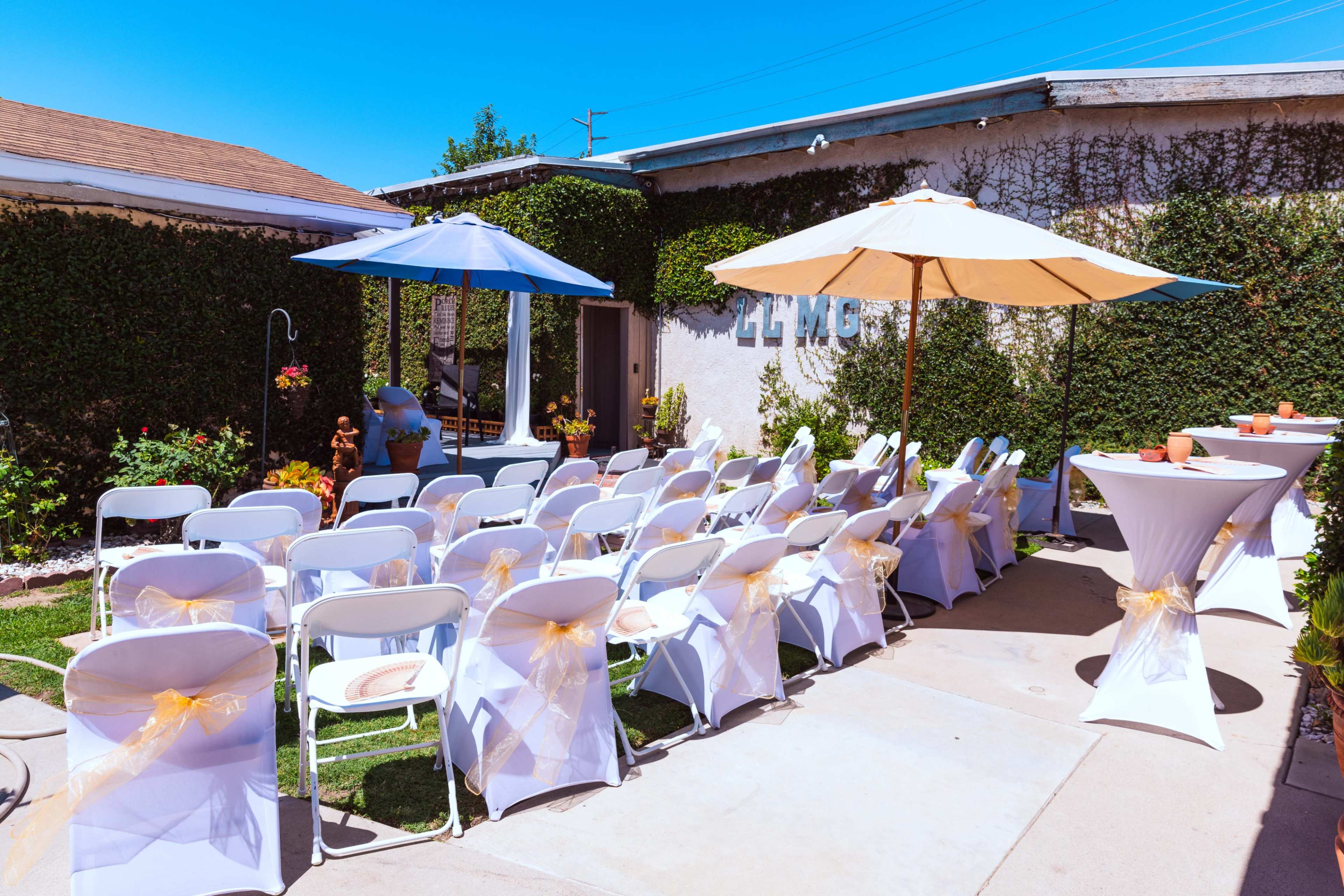 The image displays an outdoor event setup with rows of white chairs adorned with yellow sashes, two umbrellas providing shade, and a table set for refreshments.