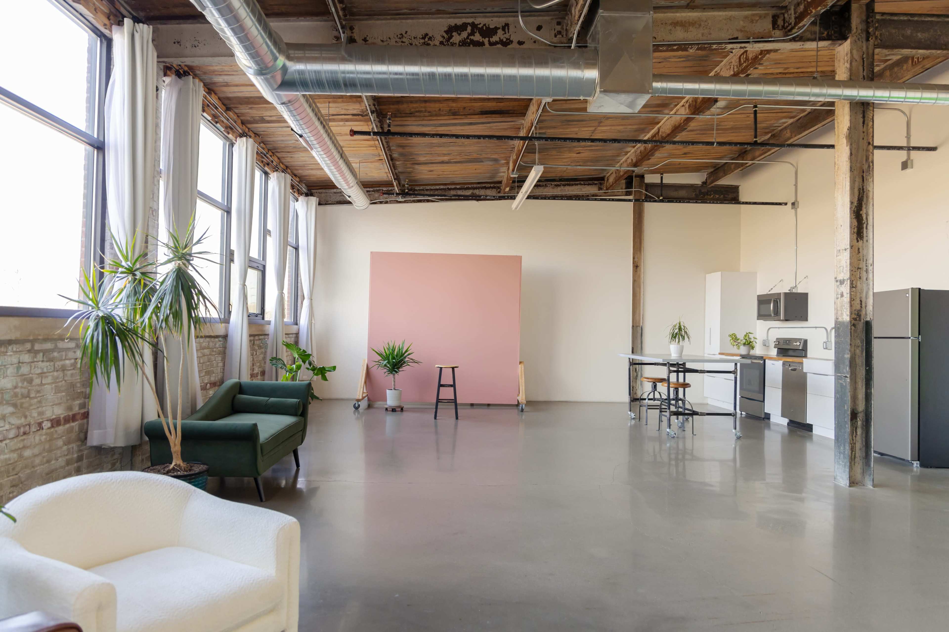 A spacious, industrial-style room with large windows, concrete floors, and various plants, featuring a pink backdrop and kitchen appliances along one wall.